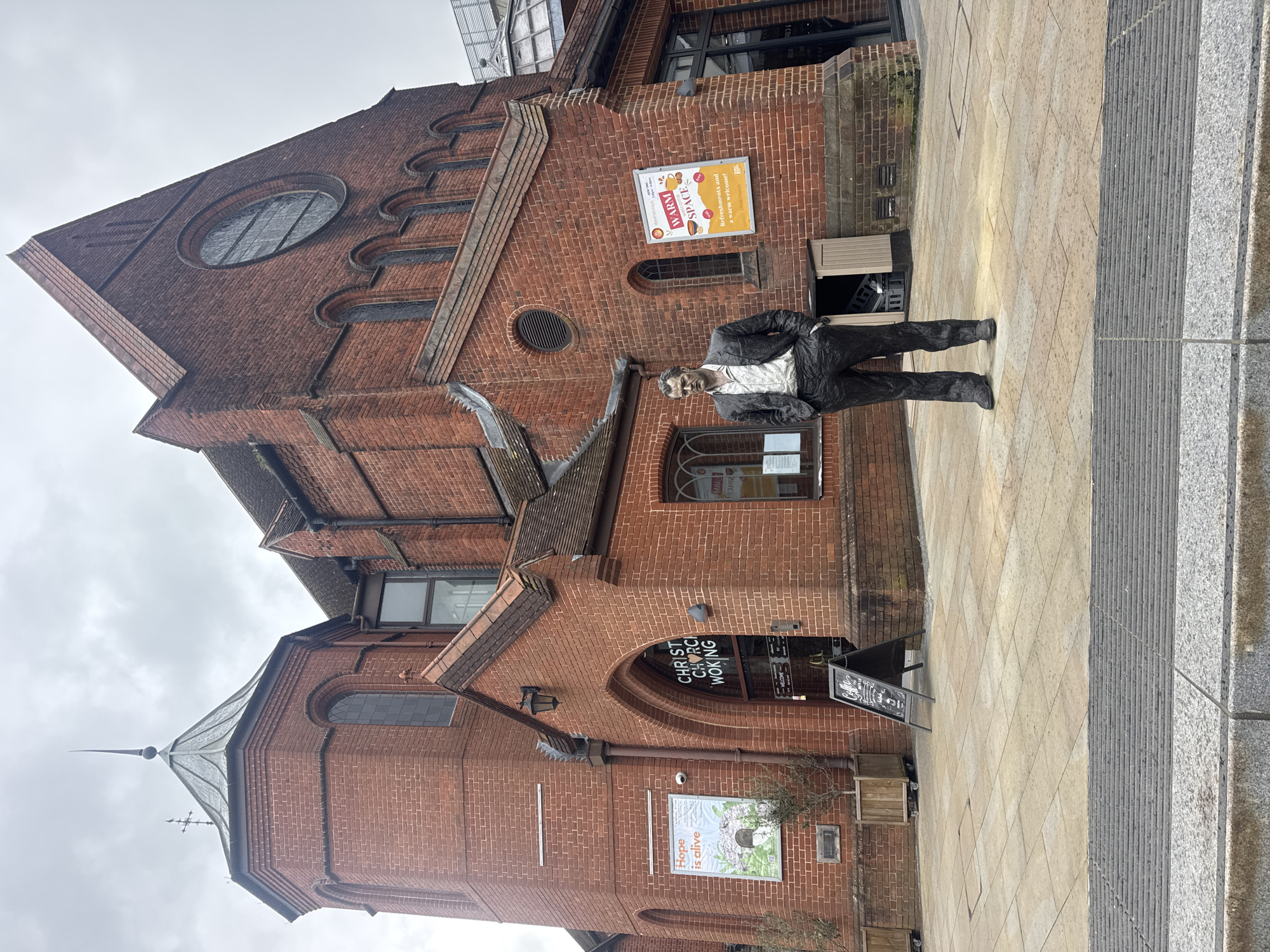 This image captures the exterior of Christ Church Woking, a red-brick ecclesiastical building located in Woking, United Kingdom, under an overcast sky. The prominent structure features a large main section with a high-pitched, tiled roof and a significant circular stained-glass window at its apex. To the left, a multi-sided tower with arched windows culminates in a conical metallic spire topped with a finial and a weather vane incorporating a cross.
A dark-colored, life-sized statue of a man stands on the light-colored paved forecourt directly in front of the church. The man is depicted wearing a suit jacket, an open-collared shirt, and trousers, with his hands placed in his pockets. The statue has realistic features and is positioned facing slightly right from the viewer's perspective.
The church's entrance area, beneath an arched red-brick canopy, features glass doors displaying "CHRIST CHURCH WOKING." To the left of the entrance, a poster titled "Hope is alive" is mounted on the wall. To the right of the entrance, another poster advertises a "WARM SPACE" offering "Refreshments and a warm welcome! free," attributed to Christ Church. An A-frame chalkboard sign, partially visible near the entrance, advertises a "Coffee Shop."
The immediate ground consists of uniformly laid square paving slabs in a light tan hue. A dark grey, horizontally ridged tactile paving strip is visible in the lower foreground, bordering a darker grey curb. Wooden planters containing small, bare-branched trees are situated along the church wall to the left of the entrance. No human subjects or active interactions are visible in the scene.