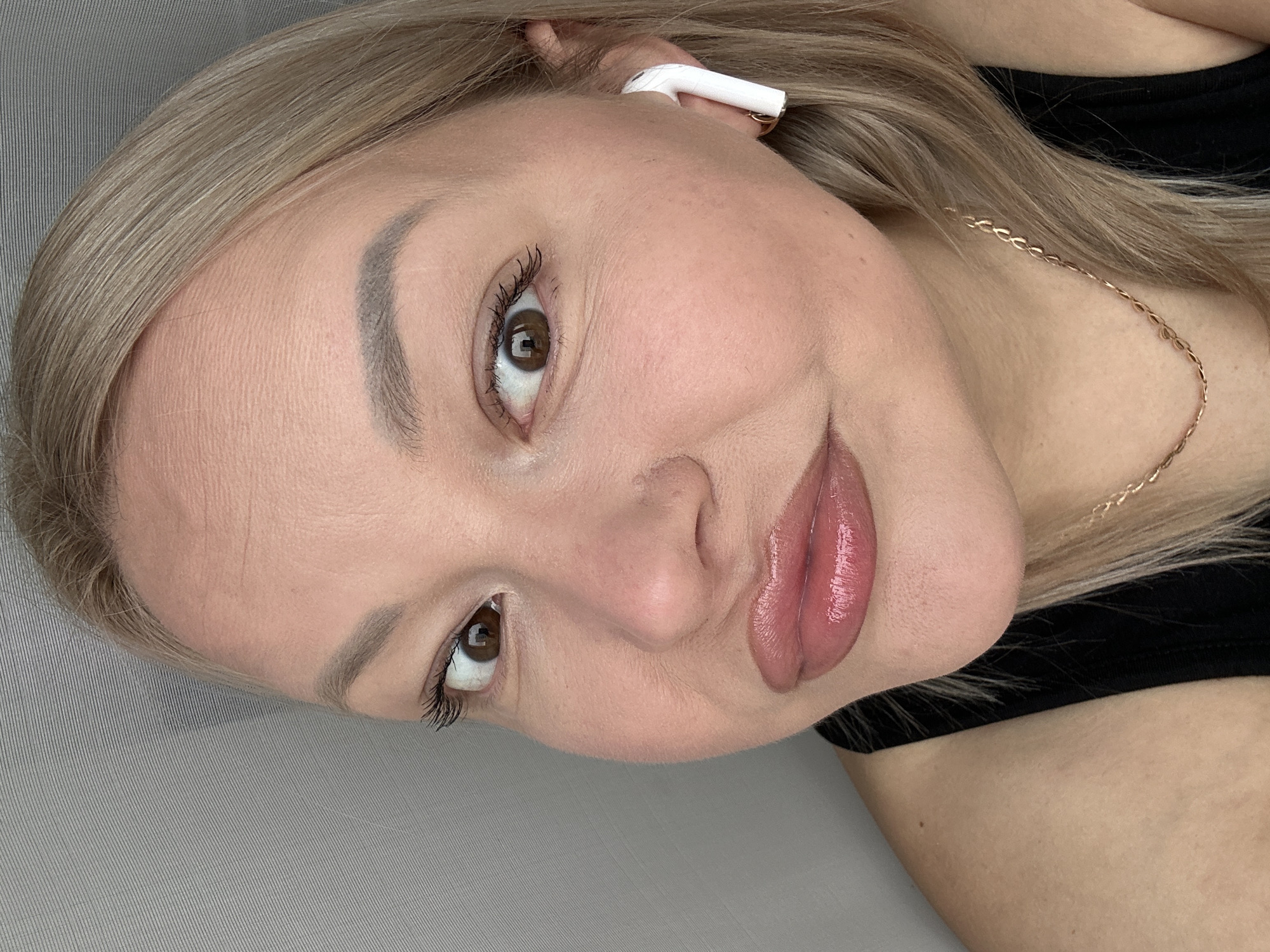 A close-up, angled portrait photograph of a woman with light blonde hair, fair skin, and brown eyes. She is wearing a white earbud in her left ear and a delicate gold chain necklace. She has full lips with a glossy, pinkish color. The background is a soft, neutral grey. There is no information available that connects this image to the city of Izhevsk, Russia.