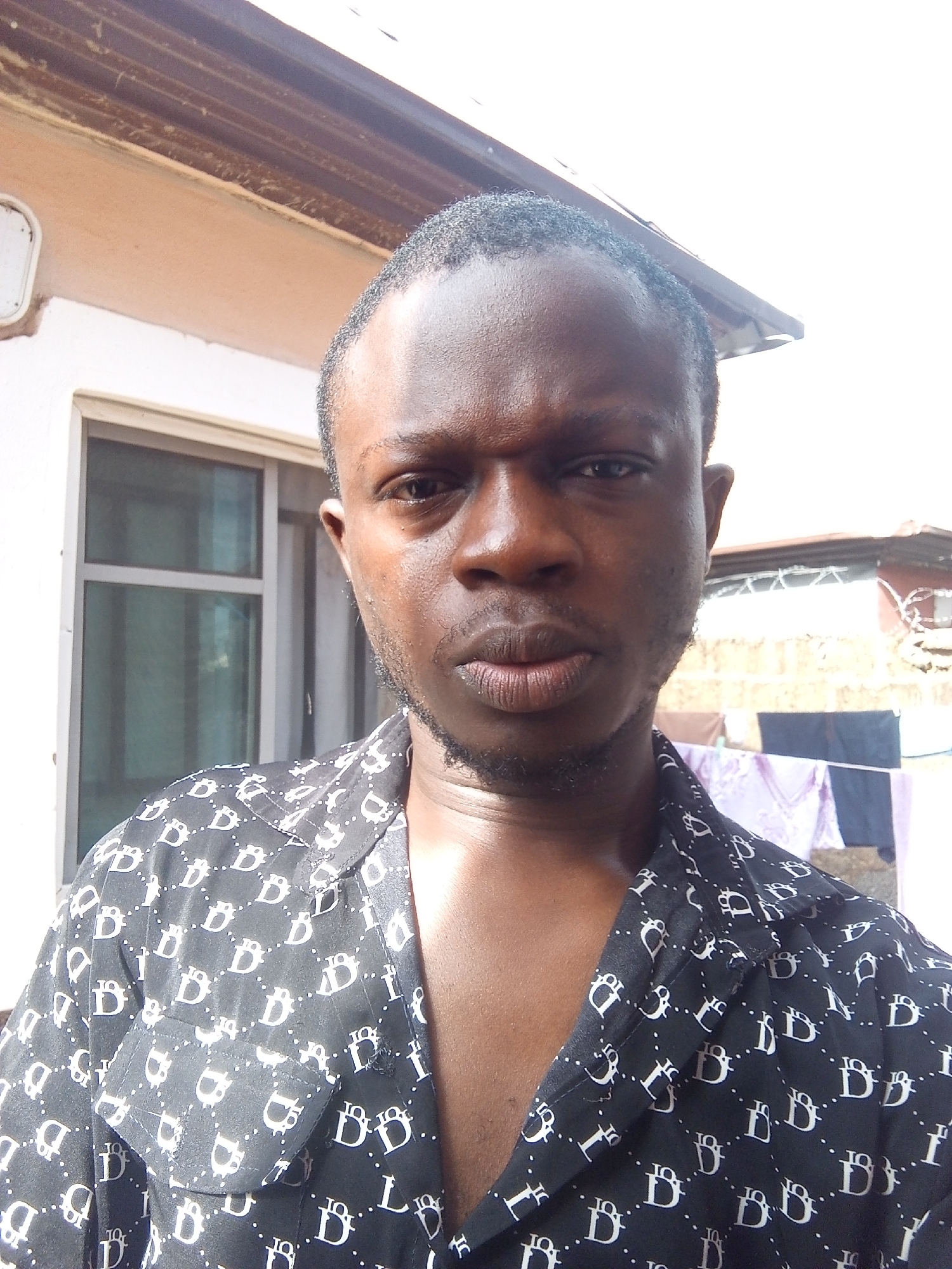 A medium close-up, eye-level, outdoor, selfie-style photograph shows a Black man with short, dark hair, a light beard, and light skin. He is wearing a black short-sleeved button-up shirt with a repeating white logo pattern. He is looking directly at the camera with a serious expression. His head and shoulders are visible, and he is positioned slightly off-center to the right.
In the background, there is a building with light-colored walls and a brown, corrugated metal roof overhang. To the left of the man's head is a white rectangular light fixture attached to the building. Behind the man and to the right, a window with translucent panes is visible. Further in the background, laundry is hanging on a line, with purple and dark blue items visible. The overall environment suggests a residential area, possibly in a city like Abuja, Nigeria, given the architectural style and outdoor setting. The lighting is bright, indicating daytime.