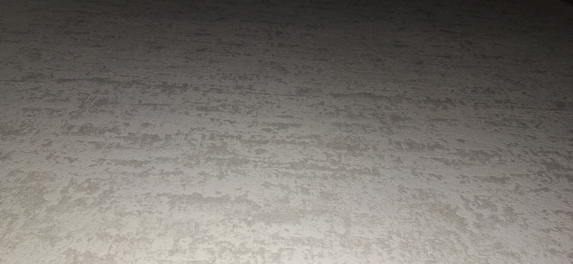 This is a full-frame, high-angle, medium shot of a textured wall. The lighting is coming from the left, creating a gradient of light and shadow across the surface. The wall has an irregular, rough texture with vertical streaks and patches of varying shades of gray and white, resembling a brushed or stucco finish. There are no people, objects, or any other discernible elements in the image. The scene is static and shows only the wall. The location in Bila Tserkva, Ukraine, is not visible or inferable from the image itself.
