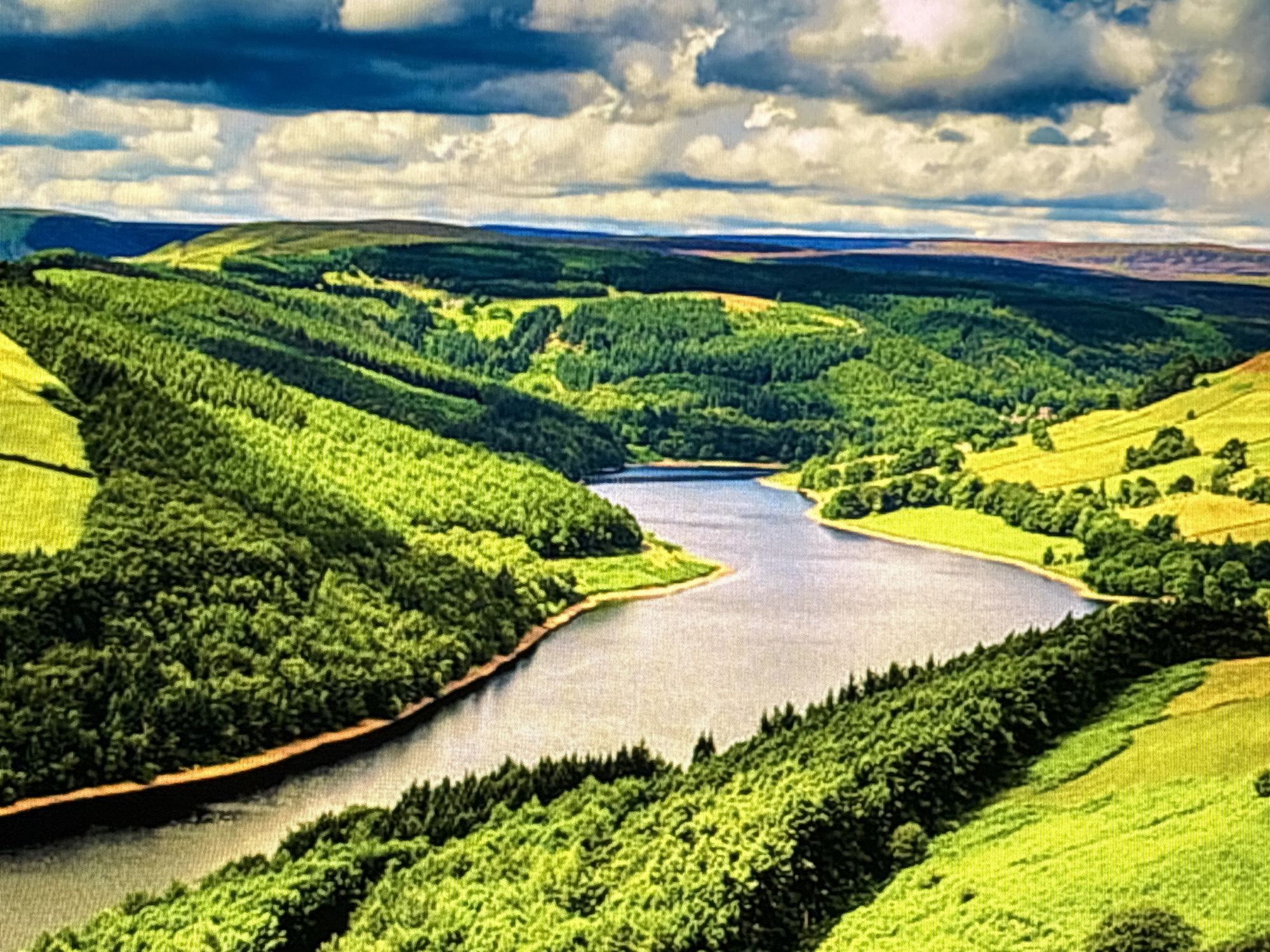 The image depicts a wide, scenic aerial view of a body of water, likely a reservoir, winding through rolling green hills covered in dense forests and grassy meadows. The sky above is dramatic, with dark, heavy clouds interspersed with patches of bright white and blue. The landscape is characterized by lush vegetation, with distinct contrasts between the deep green of the forests and the lighter, yellow-green of the sunlit meadows. The water is a calm, slate gray, reflecting the sky in places. In the distance, a distant ridge line forms the horizon, appearing as a dark silhouette against the sky. There are no people or man-made structures immediately visible within the frame, giving the impression of a natural, untouched landscape.
This image does not depict any location in the city of Sofia, Bulgaria. The scene is characteristic of a mountainous or hilly region with significant forestation and water features, such as national parks or reservoir areas.