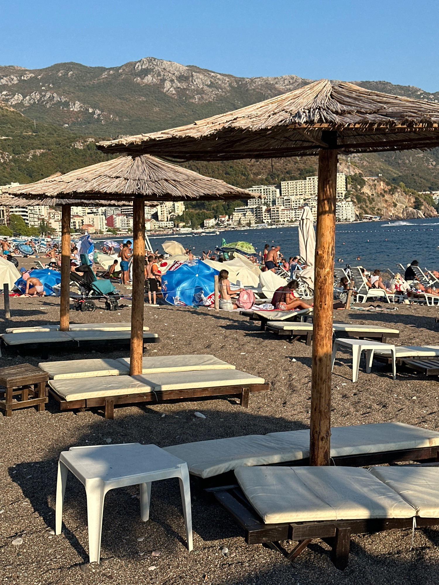 In Boreti, Montenegro, a sun-drenched beach scene unfolds under a clear blue sky. The foreground features several empty, dark wooden lounge chairs with light-colored cushions, arranged on a pebbly shore. Two straw-thatched umbrellas, supported by thick wooden poles, provide shade. In the middle ground, a bustling beach is populated by numerous sunbathers. People are seen relaxing on lounge chairs, sitting on towels, and wading in the calm, blue sea. A stroller is visible among the beachgoers. In the background, a dense cluster of white buildings, likely hotels or apartments, ascends the rocky, tree-covered hillside. The distant mountains, with their rugged, rocky peaks, form a dramatic backdrop. The overall impression is one of a lively summer day at a popular coastal resort.