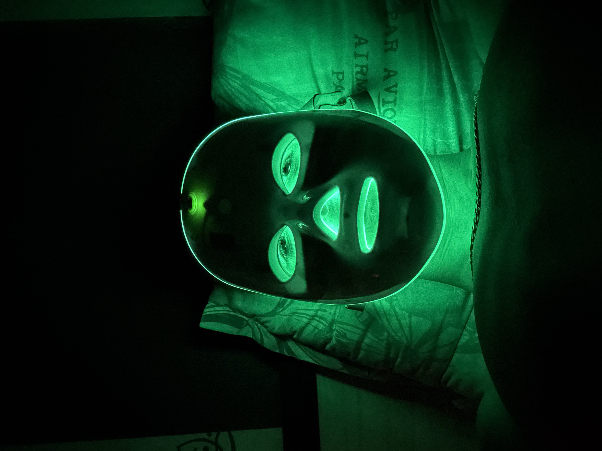 The image shows a person wearing a green LED facial mask, lying on a bed or couch. The mask emits a bright green light, illuminating the person's face and neck. The background is dark, with some soft lighting visible on the right side. Text on a pillow or clothing behind the person is partially visible, reading "PAR AVION AIRMAIL". The scene is indoors, and the overall impression is one of a nighttime or dimly lit environment. No specific location in Stara Zagora, Bulgaria, can be identified from the image.
