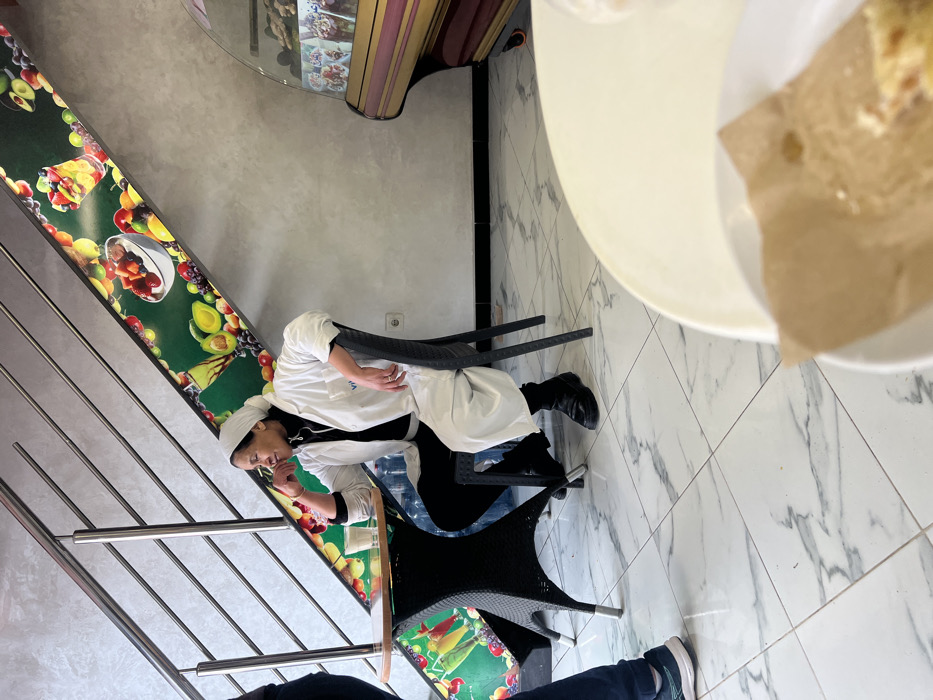An adult female, identified as an employee or owner, is seated at a small round table within a commercial establishment in Meknes, Morocco. She is positioned towards the left-center of the frame, wearing a white headscarf, a white lab coat over a black turtleneck, and black trousers with black boots. Her left hand rests near her mouth as she looks towards the left, out of frame. A clear glass containing a light-colored beverage is on the table in front of her. To her immediate left, a portion of another person, clad in blue jeans and white and blue sneakers, is visible, standing near the table.
The floor is covered with large, polished white tiles featuring gray marble-like veining. The walls are a textured light gray. A modern staircase with stainless steel railings ascends diagonally from the left foreground to the upper right background. The wall section beneath the staircase is adorned with a vibrant green mural or decal depicting various fruits like avocados, strawberries, grapes, and limes, alongside images of fruit salads and smoothies.
On the right side of the image, a curved, glass-fronted display case with a dark red or burgundy base is present, showcasing what appear to be pastries or other food items. A white electrical outlet is visible on the lower wall behind this display case. In the foreground, partially visible on the right, is a section of a large, round, white table, on which a white plate holds a brown paper-wrapped item, likely a pastry or snack. Several large, clear water bottles are positioned on the floor beneath the table where the main subject is seated. The scene suggests a quiet moment in a cafe or snack bar.