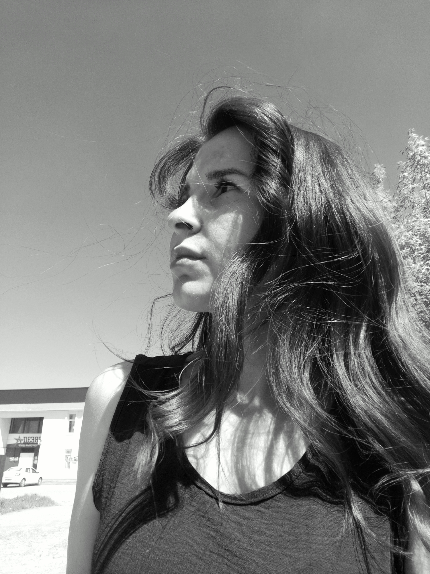 A black and white medium shot depicts a woman with long, wavy hair looking toward the upper left. The wind is blowing her hair, and some strands are across her face. She is wearing a dark tank top. The background features a building with the Cyrillic script "А-ДЕЗ-К" visible, indicating a possible address or business name. A light-colored car is parked in front of the building. The scene appears to be outdoors in a city, likely in Фоки, Russia, given the script on the building. A tree is visible in the upper right corner. The sky is clear.