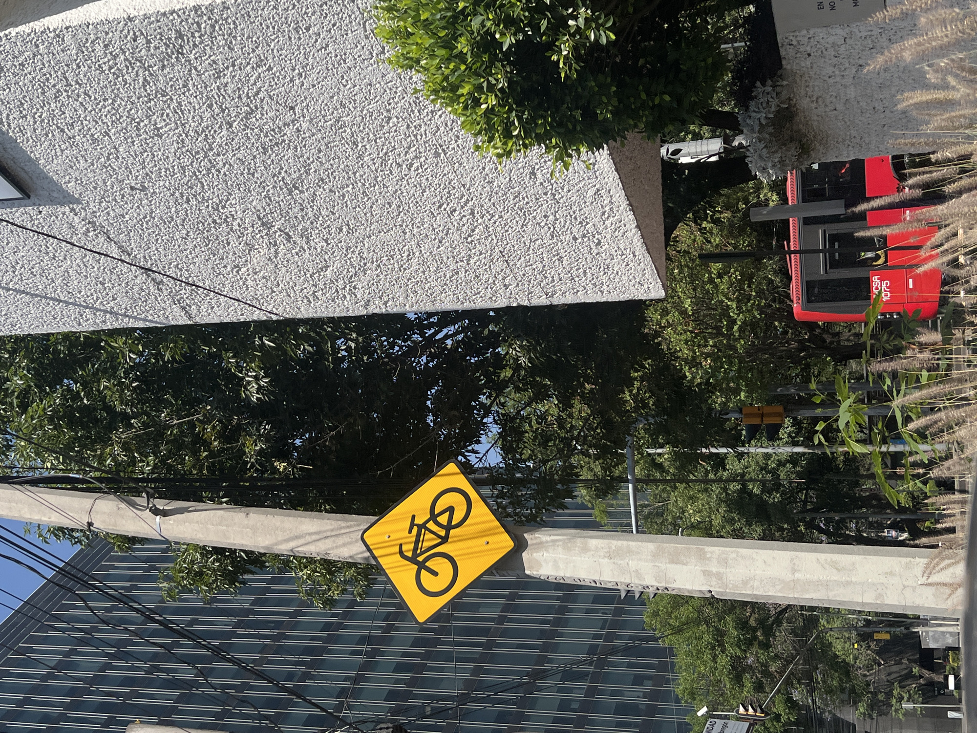 The image captures a sunny urban street scene in Mexico City, showcasing a mix of infrastructure, buildings, and greenery. The original image is rotated 90 degrees clockwise, but when viewed upright, the scene depicts a lively cityscape under clear skies.
In the mid-ground, a bright yellow, diamond-shaped sign featuring a black bicycle icon is prominently displayed, indicating a cycling path or area. This sign is mounted on a sturdy concrete utility pole that runs vertically through the left side of the frame, carrying multiple black power lines.
To the left of the pole, a large, modern high-rise building with a dark, reflective glass facade and horizontal white banding fills the background, reflecting the bright blue sky. On the right side of the frame, a red public transport bus is visible, its modern design and dark windows suggesting a city bus system. The text "VCSA 1075" is clearly visible on its side. Above and behind the bus, a building with a textured, light-colored stucco-like finish can be seen.
Lush green trees are scattered throughout the scene, providing natural elements and casting shadows, indicative of bright daylight. Some ornamental grasses and plants are also visible along the edges of the frame, particularly near the bus. The presence of traffic lights (yellow components visible) further down the street confirms an active urban environment. No specific people or activities are central to the frame, but the elements suggest a typical day in a bustling city. Other partial text fragments visible include "EN", "NO", "M" on a white sign near the bus, and "NIDAD", "aragon" on a sign further below.