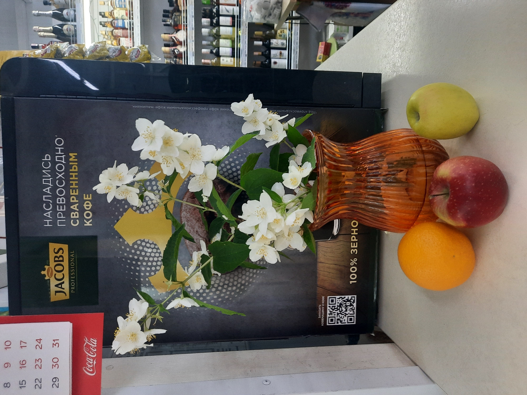 In the city of Porechye, Belarus, a still life composition is set against the backdrop of a coffee machine advertisement. The advertisement features the Jacobs Professional coffee brand logo and text in Russian, with a QR code and the phrase "100% ZERNO" (100% grain) visible. A bouquet of white jasmine flowers in a fluted orange glass vase is prominently displayed in front of the advertisement. Below the vase and flowers, three pieces of fruit are arranged: a red apple, an orange, and a yellow apple. To the right of the coffee machine, shelves stocked with bottled beverages, likely alcohol, are visible. The scene appears to be indoors, possibly in a store or cafe, with a counter in the foreground where the fruit and vase are placed. A calendar showing the month of March with dates visible is to the left of the advertisement, and a Coca-Cola logo is also present in the upper left corner.