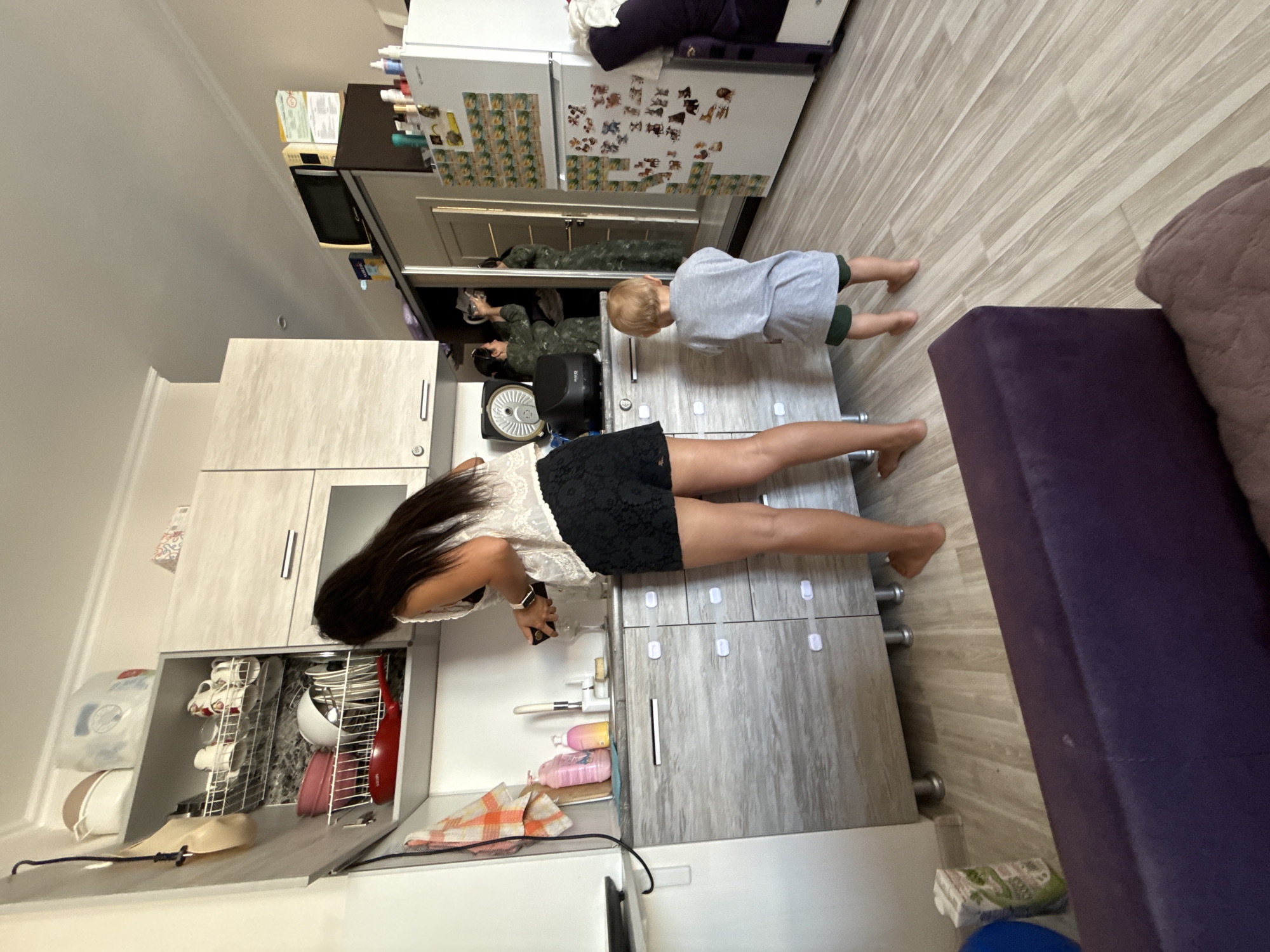 In a residential kitchen in Sochi, Russia, a woman with long dark hair, wearing a white lace top and black shorts, stands at a counter, engaged in what appears to be preparing food or drink. She holds a wine glass in her right hand and a dark object, possibly a bottle, in her left. To her right, a young child, a boy wearing a gray t-shirt and dark shorts, stands barefoot, facing away from the camera and toward a large mirror-fronted wardrobe. In the mirror, a reflection of the woman is visible, wearing a camouflage-patterned outfit and reaching into an open cabinet. The kitchen is equipped with light-colored cabinetry, a sink, and various kitchen appliances. A white refrigerator is decorated with numerous colorful stickers. The floor is made of light-colored laminate. The overall scene depicts a domestic environment with elements of everyday life.