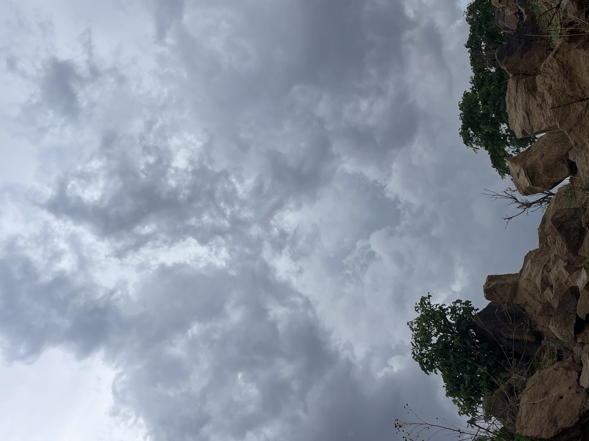 The image depicts a cloudy sky with rock formations and sparse vegetation in the upper right corner. The location cannot be confirmed as Jos, Nigeria from the visual data provided. The sky is predominantly grey, filled with layered clouds. The rock formation appears to be made of light brown stones, with a few small patches of green vegetation growing on top. Some dry, brown vegetation hangs down from the rocks.