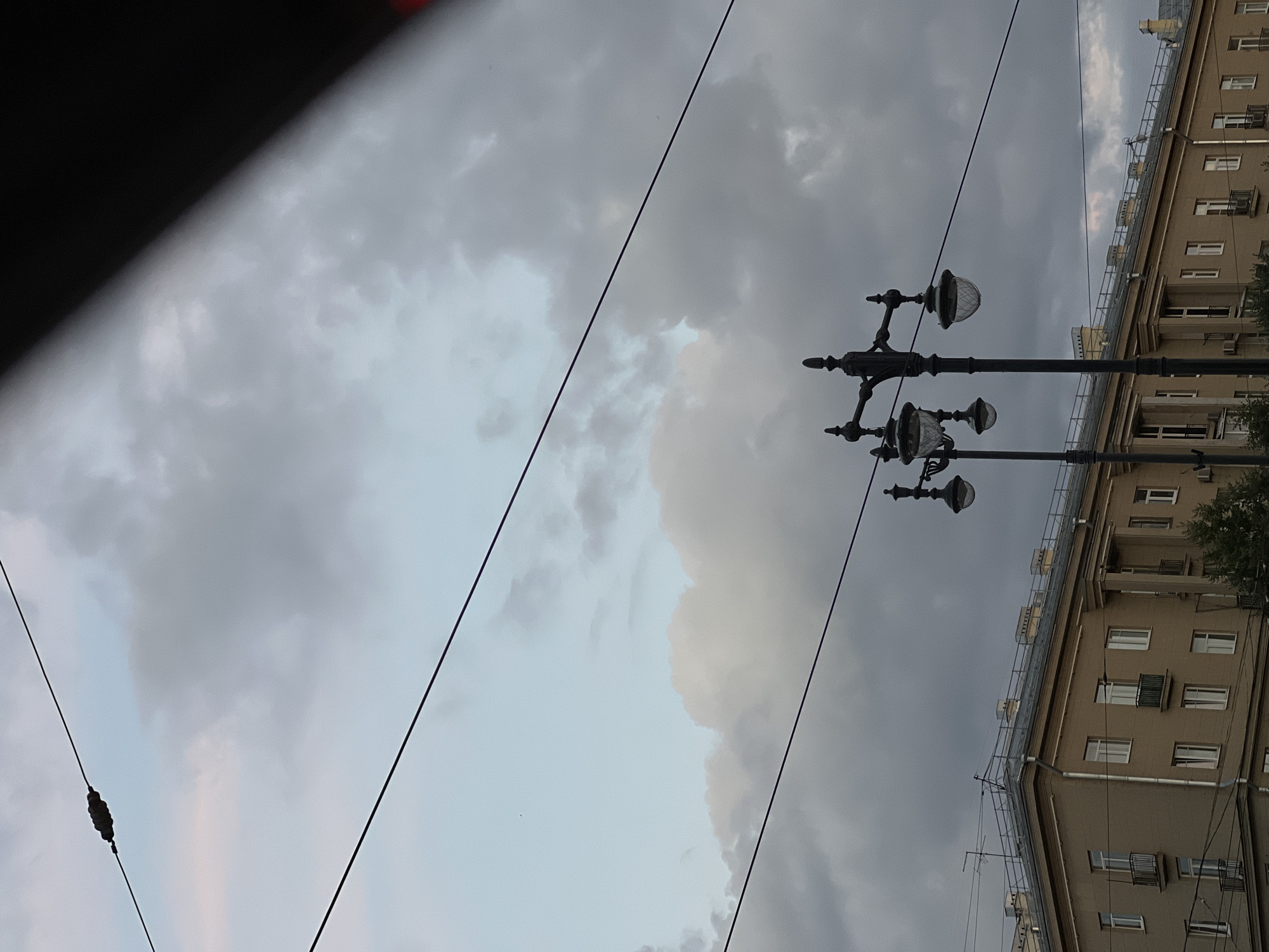 The image shows a view of a building and streetlights in Saint Petersburg, Russia. The sky is overcast with a mix of grey and light blue clouds. Power lines are visible, crossing diagonally across the frame. In the foreground, a classic streetlamp with multiple globes stands to the right. Behind the streetlamp, a long, beige apartment building with many windows and small balconies is visible. The architectural style of the building suggests it is from the Soviet era. The overall impression is of a slightly moody, overcast day in a city setting.