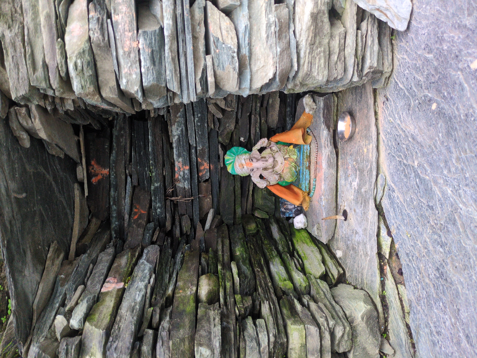 A small shrine built of stacked grey stones houses a statue of the Hindu deity Ganesha. The shrine is open at the front and has an arched ceiling formed by overlapping stones. The Ganesha statue is positioned in the center of the shrine, adorned with a green and yellow turban and an orange cloth. In front of the statue, on the stone floor, is a small metal bowl containing a reddish substance, and a short, dark, pointed object. The stones are weathered and some are covered in green moss. Red markings resembling bird tracks are visible on some of the stones inside the shrine. The surrounding environment appears to be rocky and possibly natural, with hints of vegetation. The overall scene suggests a devotional space possibly located in a natural setting or a less developed area.