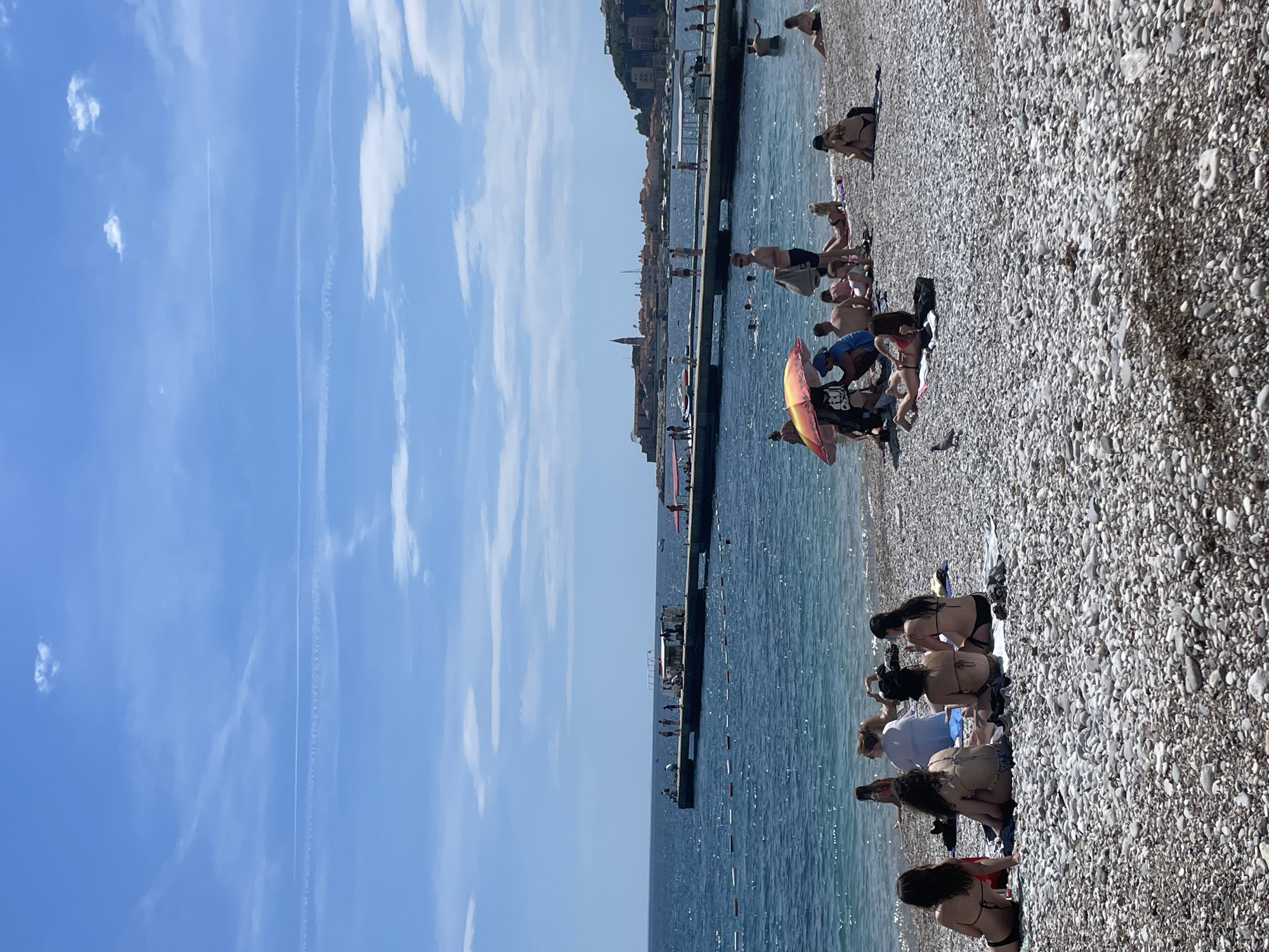 The image depicts a beach scene in Budva, Montenegro. The beach is pebbled and filled with people sunbathing and swimming. A pier extends into the water, and people are seen on the pier as well. In the background, the cityscape of Budva is visible. The sky is blue with wispy clouds.