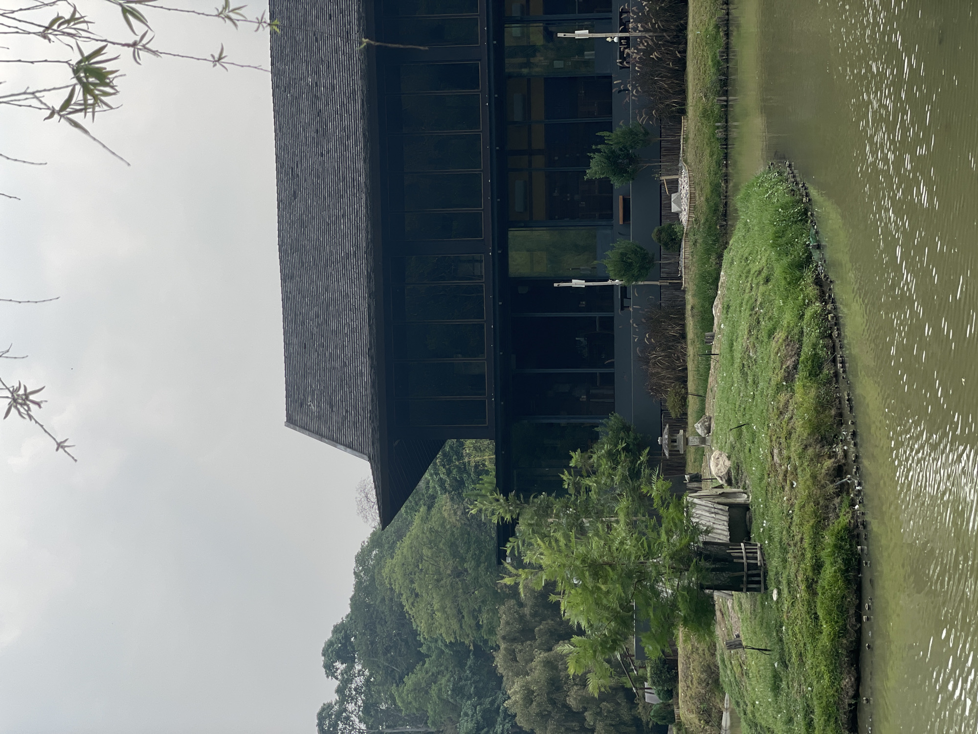 The image shows a building with a dark roof and a glass facade, situated next to a body of water. The water is a murky green color. A grassy area separates the building from the water's edge. The structure is surrounded by trees. The sky is overcast. A small wooden structure is seen on the grassy bank of the lake. The location is potentially Ban Nong Hoi, Thailand.