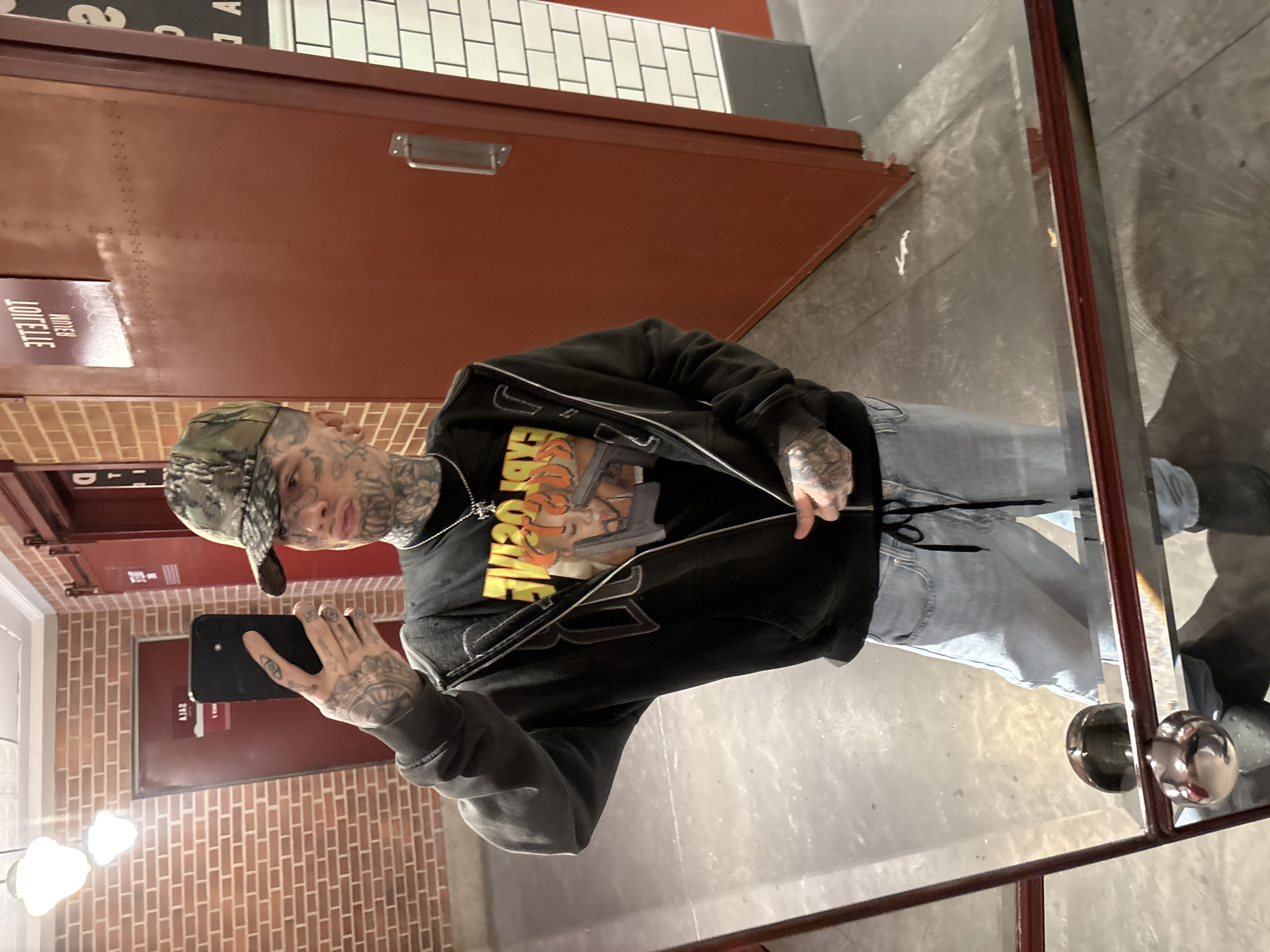 A person is captured in a mirror selfie, standing in an indoor setting that appears to be a public restroom in Buenos Aires, Argentina. The individual, who has extensive tattoos covering their face, neck, and hands, is holding a black smartphone to take the picture. They are wearing a camouflage baseball cap, a black zip-up hoodie partially open to reveal a black t-shirt, light-wash jeans, and dark shoes.
The t-shirt features a graphic print depicting a character, a handgun, and text that includes "GANGSTA" and "PARABELLUM" in a stylized, somewhat distressed font. A silver chain with a cross pendant hangs around their neck. The person's expression is neutral as they pose for the camera.
The background, reflected in the mirror, shows a wall with light-brown brick patterning on the lower half and white subway tiles above. Several brown/maroon doors are visible, one of which has a silver pull handle. Text on one of these doors is reflected and reversed, clearly reading "JOIJJUT" (TOILET) and "HITEN" (WOMEN), confirming the location as a restroom. The floor is a light, somewhat reflective surface. Two bright, indistinct white light sources are reflected in the upper left of the mirror, and two metallic, spherical door handles are visible in the reflection on the right side of the mirror frame.