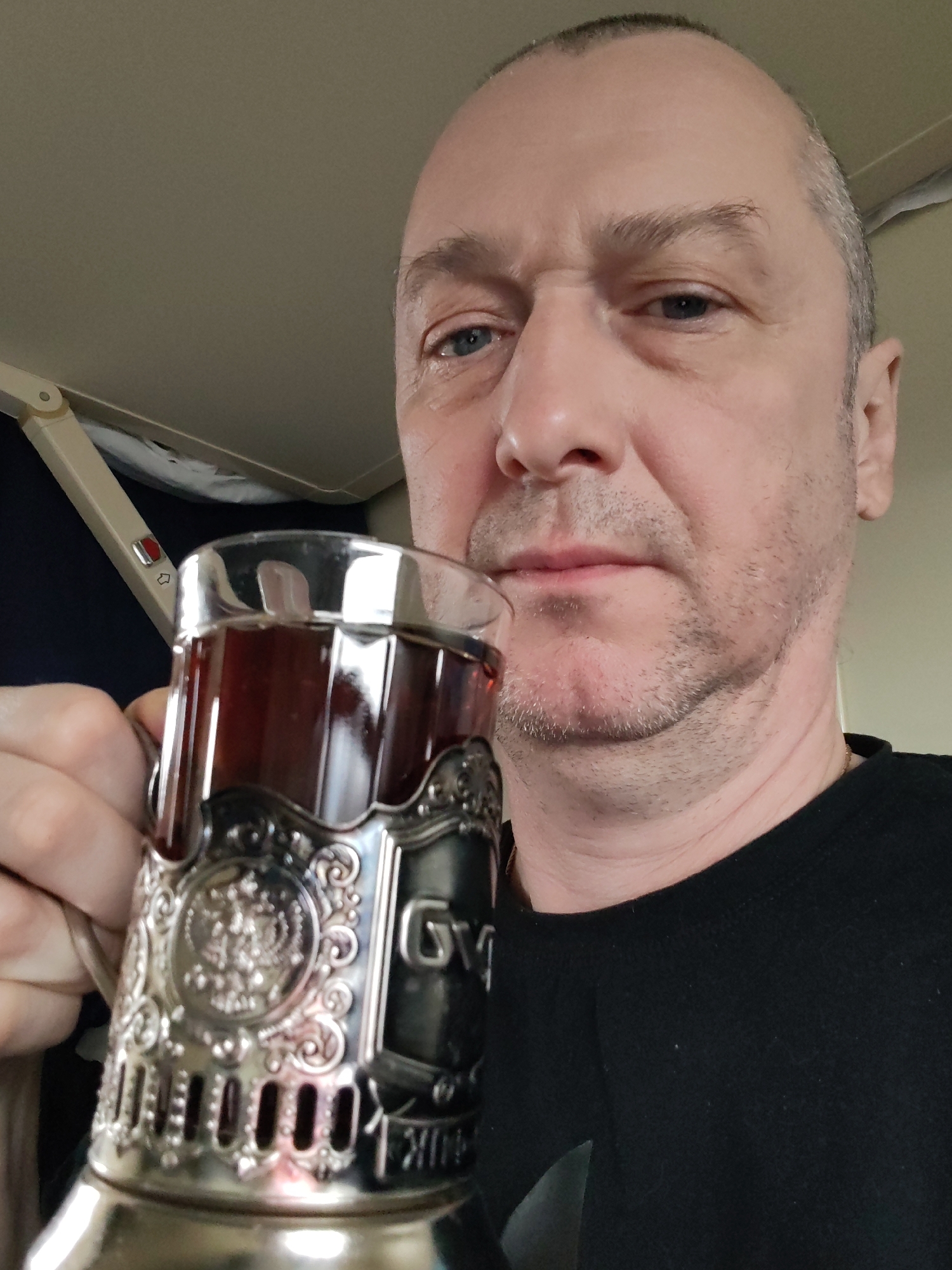 A middle-aged man with short, dark, receding hair, blue eyes, and light stubble on his face is depicted in a close-up selfie. He wears a plain black crew-neck shirt and has a neutral or slightly serious expression.
He is holding a clear glass filled with a dark liquid, likely tea, encased in an ornate silver-colored *podstakannik* (metal glass holder). The *podstakannik* features intricate embossed designs, including a prominent double-headed eagle emblem, commonly associated with Russia. Faint Cyrillic text is visible on its side, reading "СГУП" and "РЖД", with "РЖД" being the acronym for Russian Railways (Российские железные дороги).
The setting appears to be the interior of a train carriage or an airplane cabin, suggested by the light-colored overhead panel and a folded, beige-colored tray table arm with a small red button and an icon. The man is holding the glass up towards the camera, seemingly taking a picture while seated. The location context is Бордуки, Russia, which aligns with the Russian Railways branding on the *podstakannik*.