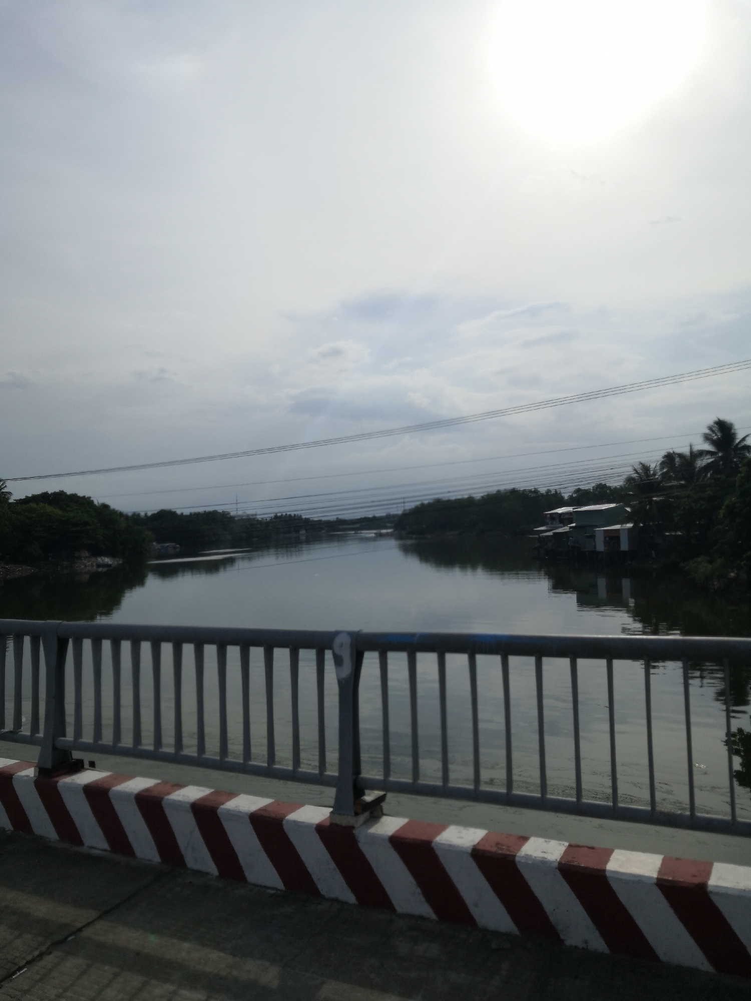 The image captures a view of a river from a bridge in Bac Nha Trang Ward, Vietnam. A grey metal railing with vertical bars runs across the foreground, separating the bridge from the river. The base of the railing is a concrete barrier painted with diagonal red and white stripes. The river, reflecting the overcast sky, stretches into the distance, lined with dense green trees on both banks. Several houses on stilts are visible on the right bank amidst the trees, and power lines are strung across the sky. The sun is partially obscured by clouds, creating a bright halo effect in the upper right portion of the frame. The overall atmosphere is calm and serene.