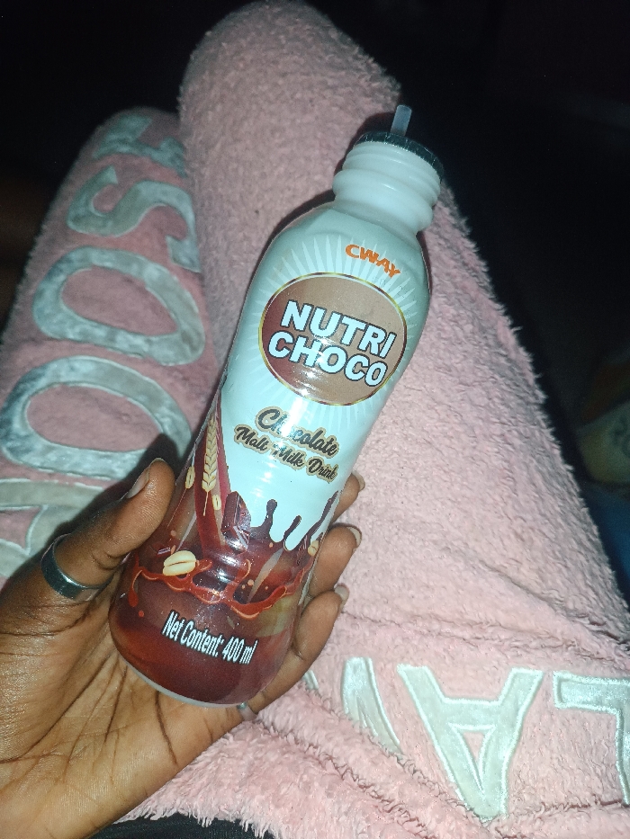 A hand holds a bottle of CWAY Nutri Choco chocolate malt milk drink. The bottle's label includes the words "Nutri Choco" and "Chocolate Malt Milk Drink" and "Net Content: 400 ml" written on the bottom. The bottle has a straw inserted into the top. The hand is adorned with a silver ring. The bottle is resting on a person's lap, clad in pink, fuzzy loungewear with the letter "L" emblazoned on it. The background is dark and dimly lit.