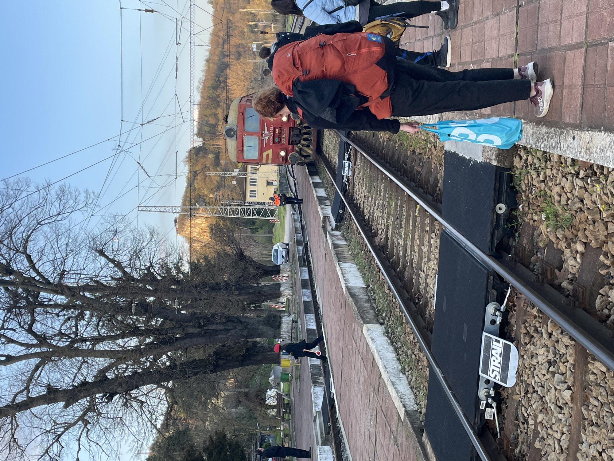 The image depicts a scene at a railway station, possibly in Veliko Tarnovo, Bulgaria. A red and yellow locomotive is approaching the platform. Several people are on the platform. One person with an orange backpack stands near the edge, holding a blue bag. Railway tracks run parallel to the platform, featuring a black STRAIL rail insulator. A railway worker in an orange vest can be seen standing on the track further down the platform. Bare trees are visible in the background. Overhead power lines are strung above the tracks.