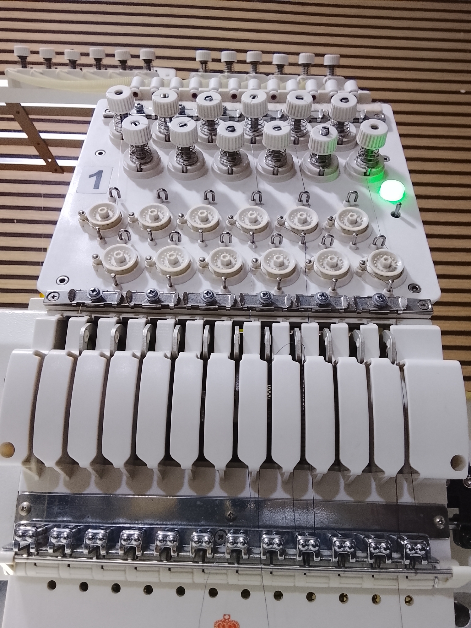 The image shows a close-up, eye-level view of an industrial embroidery machine. The machine is primarily white with metallic accents and features multiple thread spools, tensioners, and needles. In the foreground, a row of silver thread guides with individual clamping mechanisms is visible. Behind these, a series of white, rectangular components extend upwards, each housing a needle and associated thread path. Above this section, a white plate displays numerous white thread spools, each with a corresponding tension adjustment knob. One of these spools has a green light illuminated next to it. The number "1" is prominently displayed on the white plate near the spools. The background consists of horizontal wooden planks. This scene is located in a workshop or factory environment, within the city of Rumuepirikom, Nigeria.