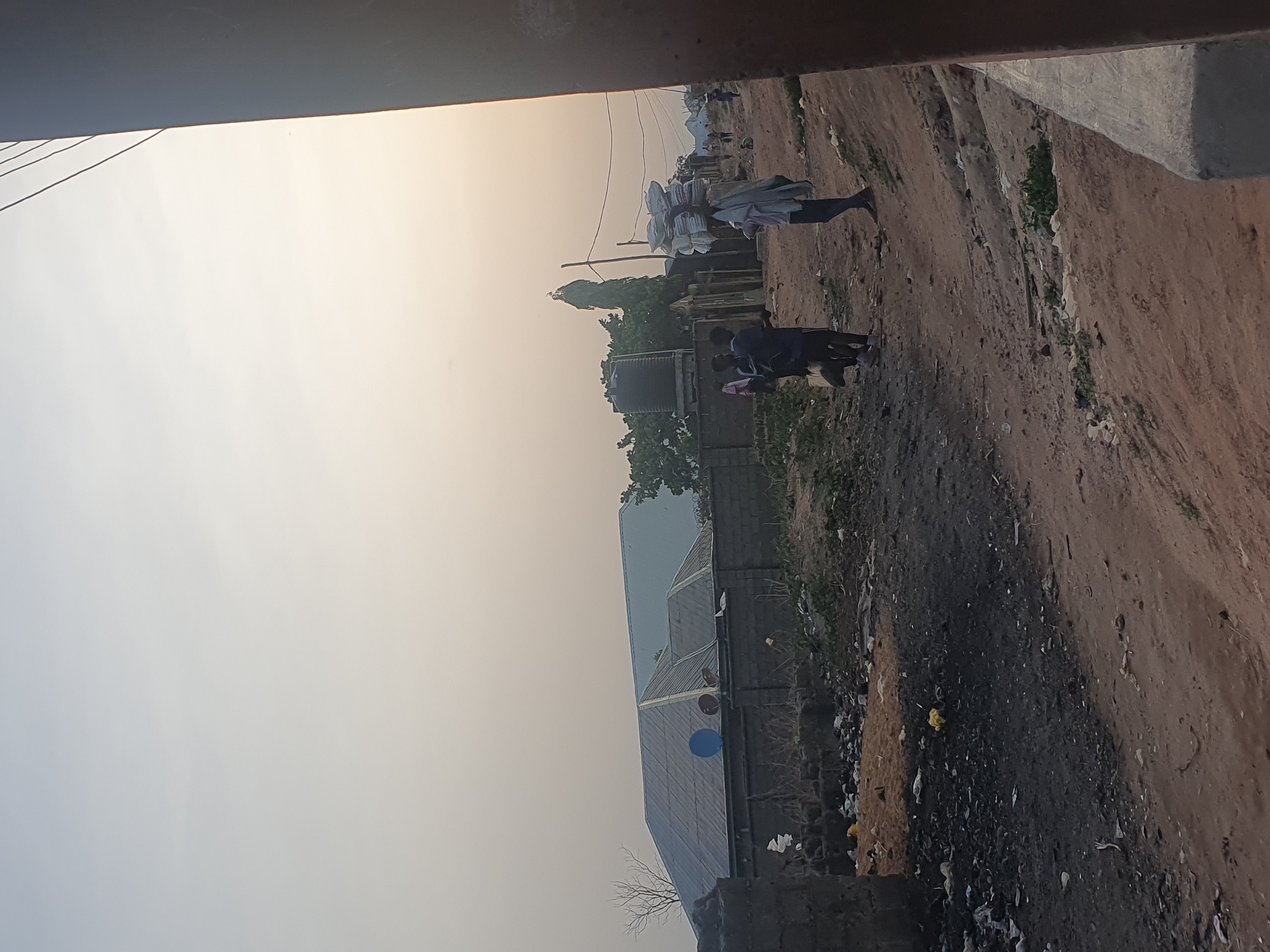 The image depicts a street scene in Lokoja, Nigeria. The dirt road is bordered by a wall and vegetation on one side and a building on the other. Two people are walking along the road. One is carrying a large load on their head. There is a water tank and a building with a corrugated metal roof visible in the background. The sky is hazy.