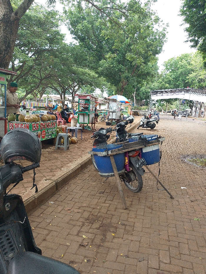 The image depicts an outdoor food stall area in Kalianda, Indonesia, under the shade of large, verdant trees. In the immediate foreground on the left, the front portion of a black scooter is visible, with a helmet resting on its handlebars. Adjacent to it, a vendor, identifiable by his green cap and light brown shirt, stands behind a stall laden with whole and opened coconuts. This stall prominently displays the text "ES DUGAN" and "ES DOCAN," indicating the sale of coconut ice beverages.
To the right of the stall, a distinctive motorcycle is parked on the brick pathway. It features a custom attachment: a large blue drum-like container secured to its rear, supported by two wooden poles propped on the ground, forming a stand. Its license plate, "BE 5720," is partially visible.
Further into the background, the paved brick pathway extends, flanked by numerous other food carts and stalls. Some of these stalls are equipped with colorful umbrellas, while others are simpler structures. Several individuals are seated at tables, suggesting they are enjoying food or drinks. More motorcycles and scooters are parked throughout the area. The scene conveys a casual daytime atmosphere, likely a local market or a public gathering space, characterized by everyday activity and natural light filtered through the extensive tree canopy.