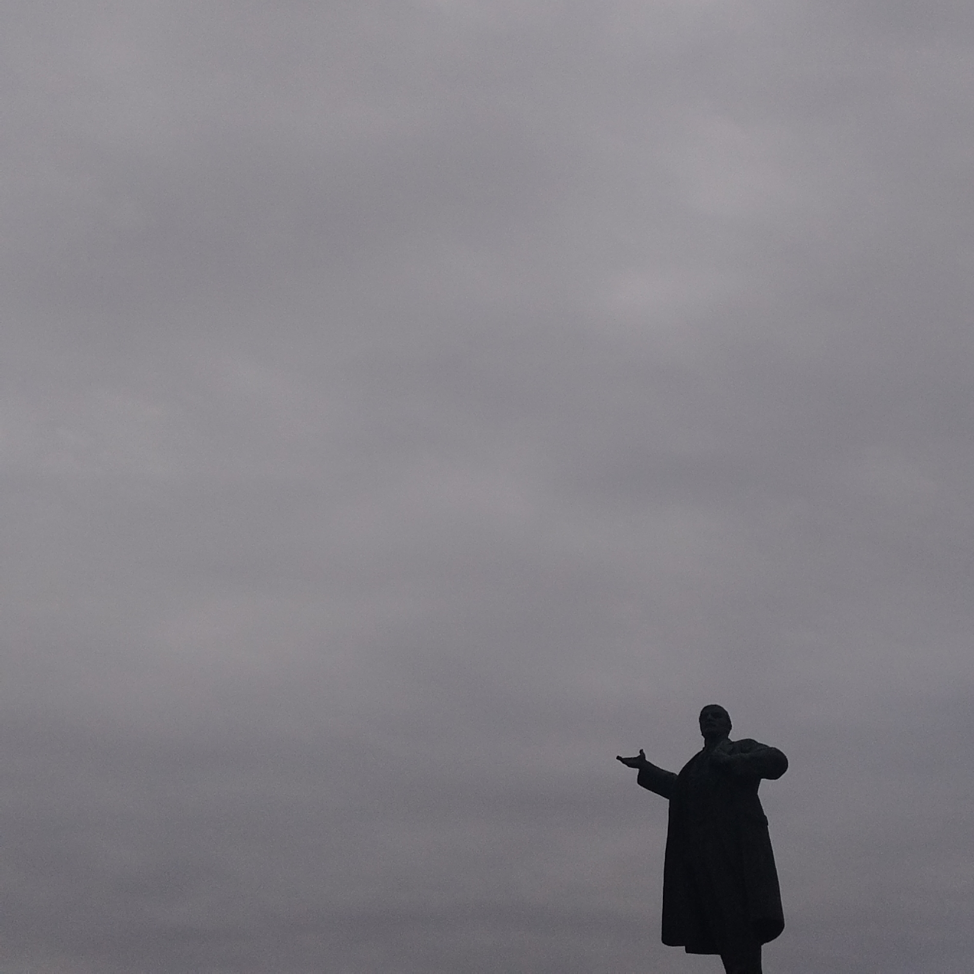 The image displays a stark, silhouetted statue of a man, likely Vladimir Lenin, against a vast, overcast grey sky. The statue is positioned in the lower right section of the frame, capturing a dynamic pose characteristic of Lenin monuments. The figure wears a long, heavy coat, and his left arm is extended forward and slightly upward, as if in a gesture of address or pointing, while his right arm is bent at the elbow and held closer to his body.
The background is entirely dominated by the uniform, dull grey of the cloudy sky, which provides a dramatic contrast to the dark silhouette of the statue. There are no other visible elements of the landscape or surrounding architecture, emphasizing the statue's isolated and monumental presence. The diffused light suggests an overcast day, contributing to a somber or contemplative mood.
Given the location context of Yekaterinburg, Russia, and the distinctive pose, the subject is clearly identifiable as a monument to Vladimir Lenin. No activity is taking place other than the static pose of the statue. There is no visible text in the media file. The overall impression is one of historical presence and solemnity under a bleak sky.