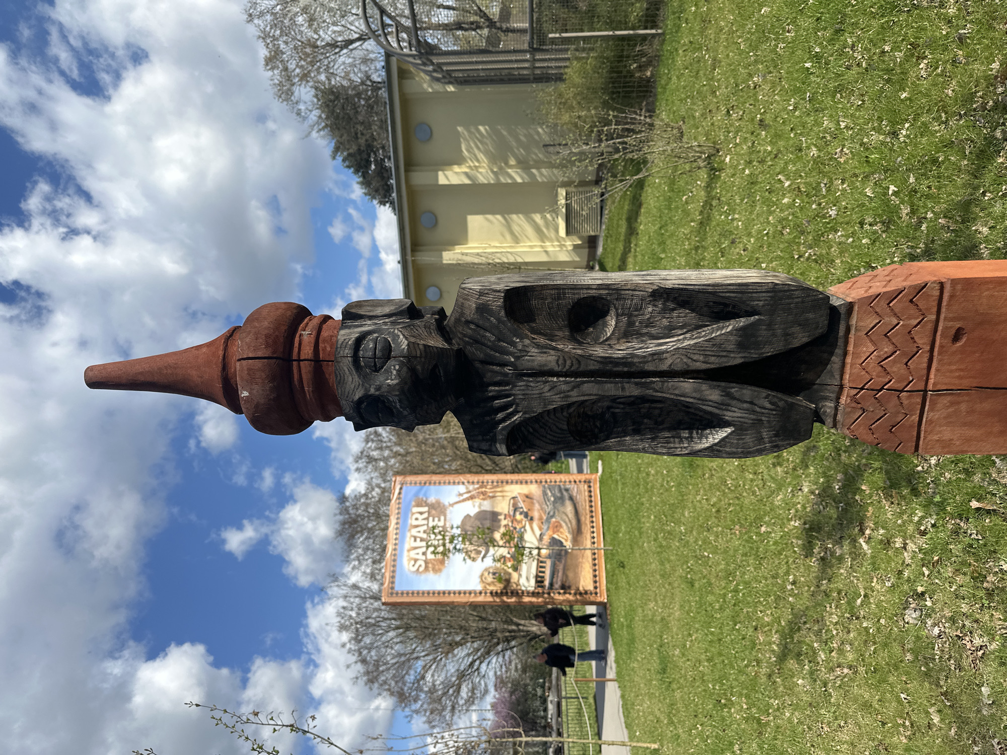 A carved wooden pole stands in a grassy area in what appears to be a park. The pole is made up of sections carved from different colors of wood. One section features a carved human head, and another section is decorated with zig-zag patterns. In the background, there is a yellow building with round windows, a "Safari" poster, trees, grass, and people. The sky is blue with white clouds. The location is likely the Wrocław Zoo in Wrocław, Poland, given the presence of the "Safari" poster and the typical architecture often found in zoos.