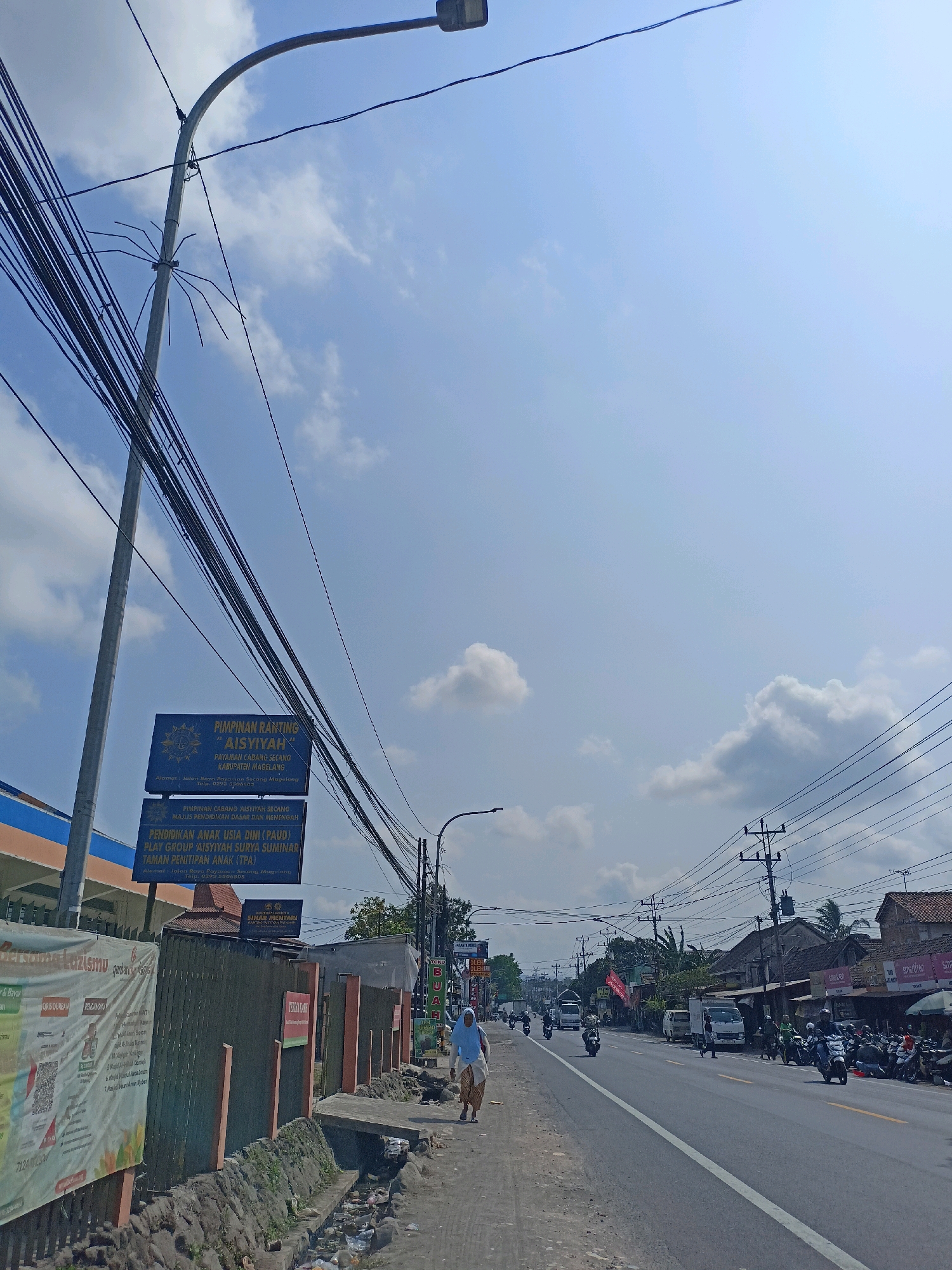 This is a street-level, eye-level, medium shot of a street scene in Payaman, Indonesia, under a bright blue sky with scattered clouds. The foreground features a paved road with a white line marking the lane. A pedestrian, wearing a light blue hijab and a tan and white patterned skirt, walks along the side of the road. To the left, there is a drain or ditch with visible debris. A low wall separates the road from a sidewalk or pathway.
Further left, a building with orange and blue accents is partially visible. A tall metal pole with numerous black wires is prominent on the left side of the image, and a decorative metal spider-like structure is attached to it. Several blue signs are mounted on a pole, one of which reads "PIMPINAN RANTING AISYIYAH" followed by information about affiliated educational institutions and an address. Adjacent to this, there is a corrugated metal fence.
The midground and background show a street lined with buildings, some with tile roofs, and trees. Traffic is visible on the road, including several motorcycles, cars, and trucks. Overhead, a complex network of utility wires and poles spans the scene. A vendor cart with a red umbrella is visible on the right side of the road, with motorcycles parked nearby. A banner on the left side of the road displays text in Indonesian. The overall impression is of a busy commercial street on a sunny day.