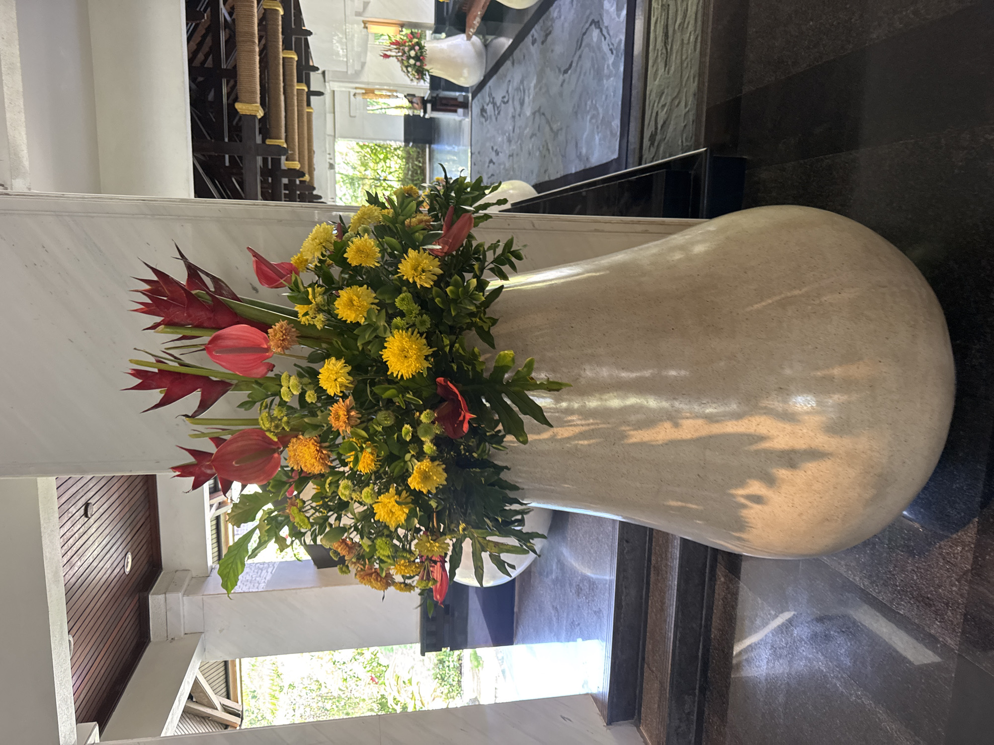 A large, beige, bulbous vase filled with a colorful floral arrangement stands on a dark, reflective floor. The arrangement features prominent red anthurium flowers, spiky red heliconia, and clusters of yellow and orange chrysanthemums, interspersed with green foliage. The vase is positioned in what appears to be a hotel lobby or a similar indoor public space, with a white marble pillar to its right and a wooden paneled ceiling visible above. In the background, through an open doorway or archway, a lush green outdoor area with trees and sunlight can be seen. Further into the building, another white vase with a smaller floral arrangement is visible on a patterned rug. The overall scene suggests a tropical resort or hotel entrance in Kuta Selatan, Indonesia, designed to create an inviting and elegant atmosphere.