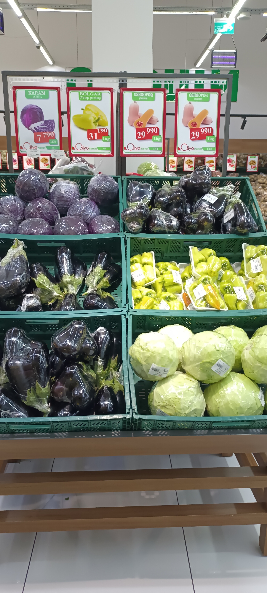 This is a full-shot, eye-level, indoor photograph of a produce display in a supermarket in Fergana City, Uzbekistan. The image captures a section of a grocery store's produce aisle, showcasing various vegetables arranged in green plastic crates. In the foreground, there are two rows of crates. The bottom row displays dark purple eggplants, each individually wrapped in clear plastic. To the right of the eggplants are several heads of green cabbage, also wrapped in clear plastic and with price labels visible. Above these, the second row of crates contains more red cabbage on the left, also wrapped in plastic. To the right of the red cabbage are more eggplants, wrapped individually. Further to the right, there are crates filled with bright green bell peppers, some in clear plastic packaging, and other vegetables like yellow bell peppers. Above the produce, promotional signs are attached to the display. One sign features a red cabbage with the price "7 790". Another sign displays yellow bell peppers with the price "31 190". A third sign shows orange squash with the price "29 990". The background shows more supermarket shelving and lighting fixtures. The environment appears clean and well-lit, typical of a modern retail setting.