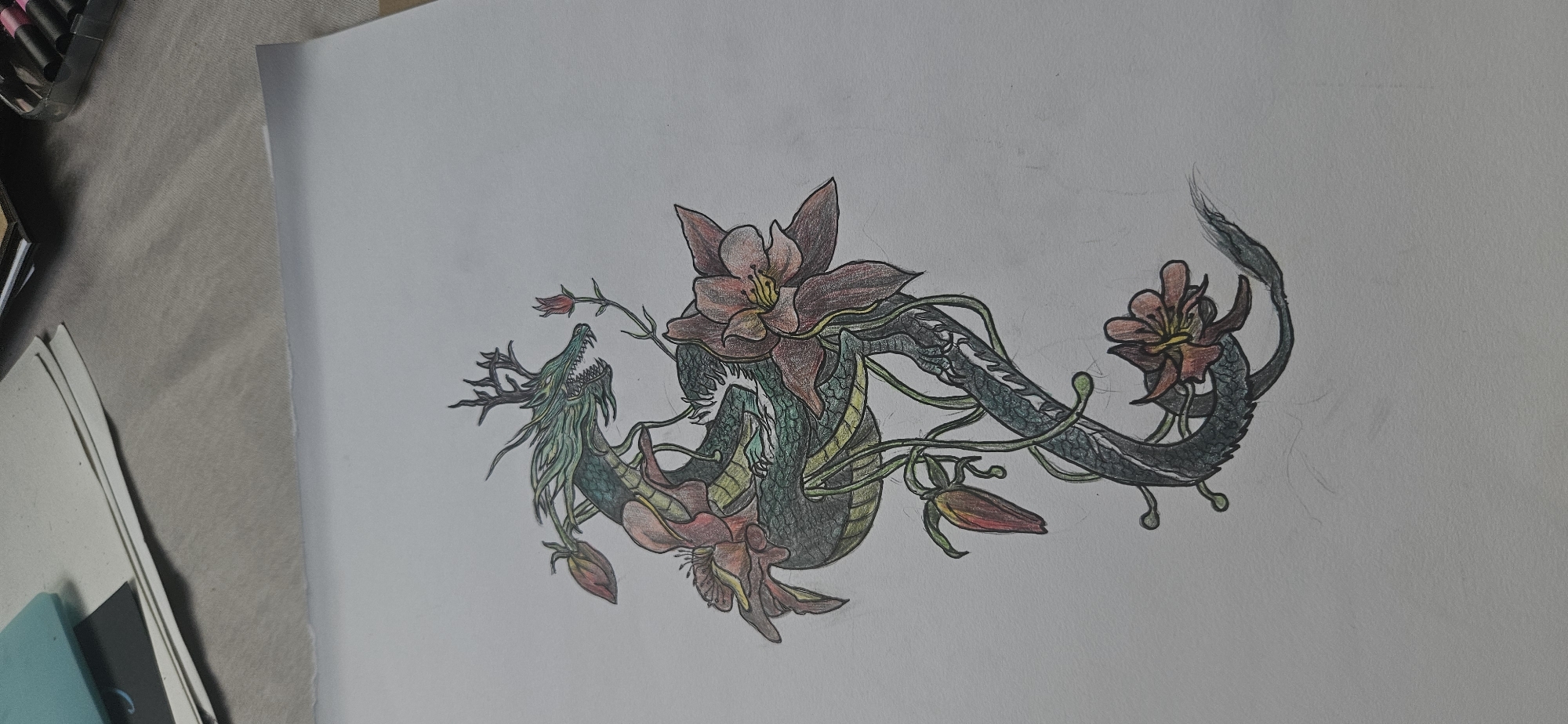 The image shows a colored pencil drawing on a white sheet of paper. The drawing depicts a stylized dragon intertwined with flowers. The dragon has a green head with antlers and an open mouth, a body with green and yellow scales, and a dark gray tail. The flowers are primarily red and pink, with green stems and leaves. The drawing is positioned in the center of the page. The background is plain white. Part of a marker set and several sheets of paper are visible in the upper left of the image. The location is not discernible from the image.