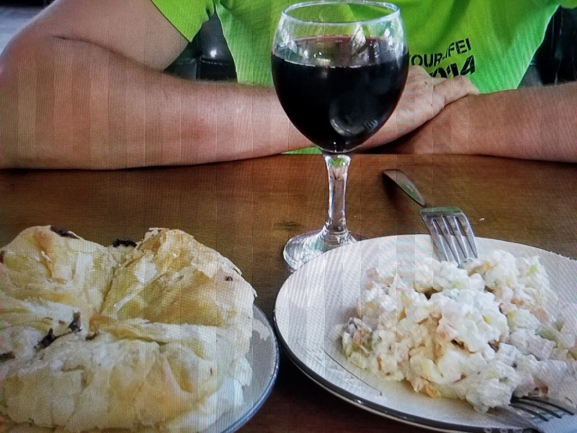 The image displays a close-up of a wooden table with two plates of food, a glass of red wine, and the arms of a person wearing a green shirt. The person's arms are resting on the table, with the left arm closer to the camera. The green shirt has some black lettering on it, partially visible.
On the left side of the image is a plate with what appears to be a flaky pastry or bread, possibly filled or topped with some dark ingredients. On the right side is another plate filled with a white salad, likely potato salad or a similar dish, with visible pieces of vegetables or other ingredients mixed in. A silver fork is placed on this plate, with its tines resting in the salad.
In the background, a wine glass filled with red liquid is positioned between the plates and the person's arms. The lighting suggests an indoor setting. The overall scene depicts a meal or snack being enjoyed at a table.
Based on the available visual information, it is not possible to determine the location within the city of Tiraspol, Moldova. The image lacks any identifiable landmarks, street signs, or architectural details that would indicate a specific place.