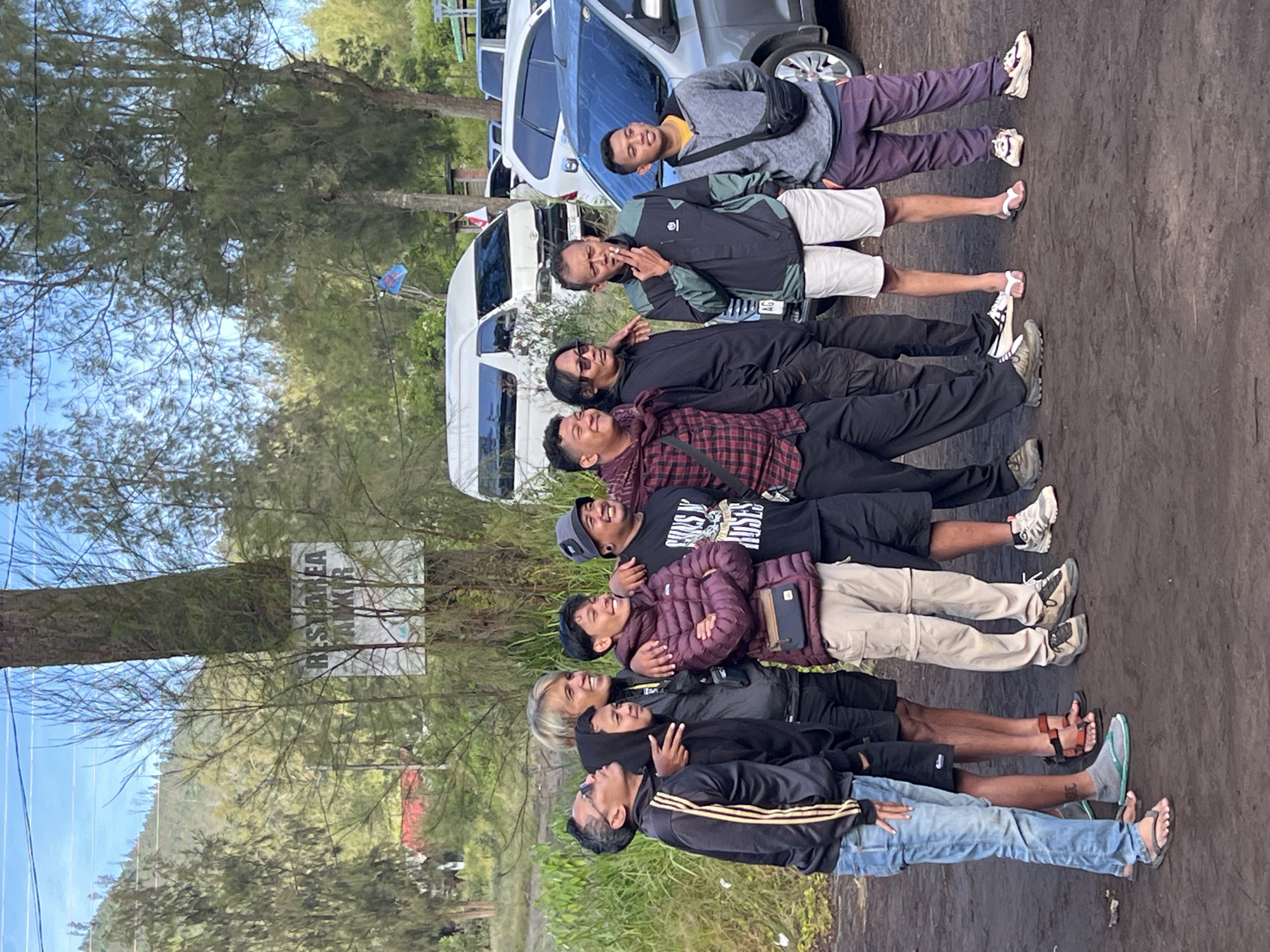 A group of eight men are lying on the ground in a row, posing for a photo. They are positioned on a dirt path, seemingly in a rural area. Trees and vegetation surround them, and several vehicles are parked nearby. A sign reading "REST AREA SANKUR" is attached to a tree. The location is likely in the city of Unknown, Indonesia. The men are wearing casual clothing, including jackets, t-shirts, shorts, pants, and sandals. They are smiling and laughing, indicating a jovial mood.