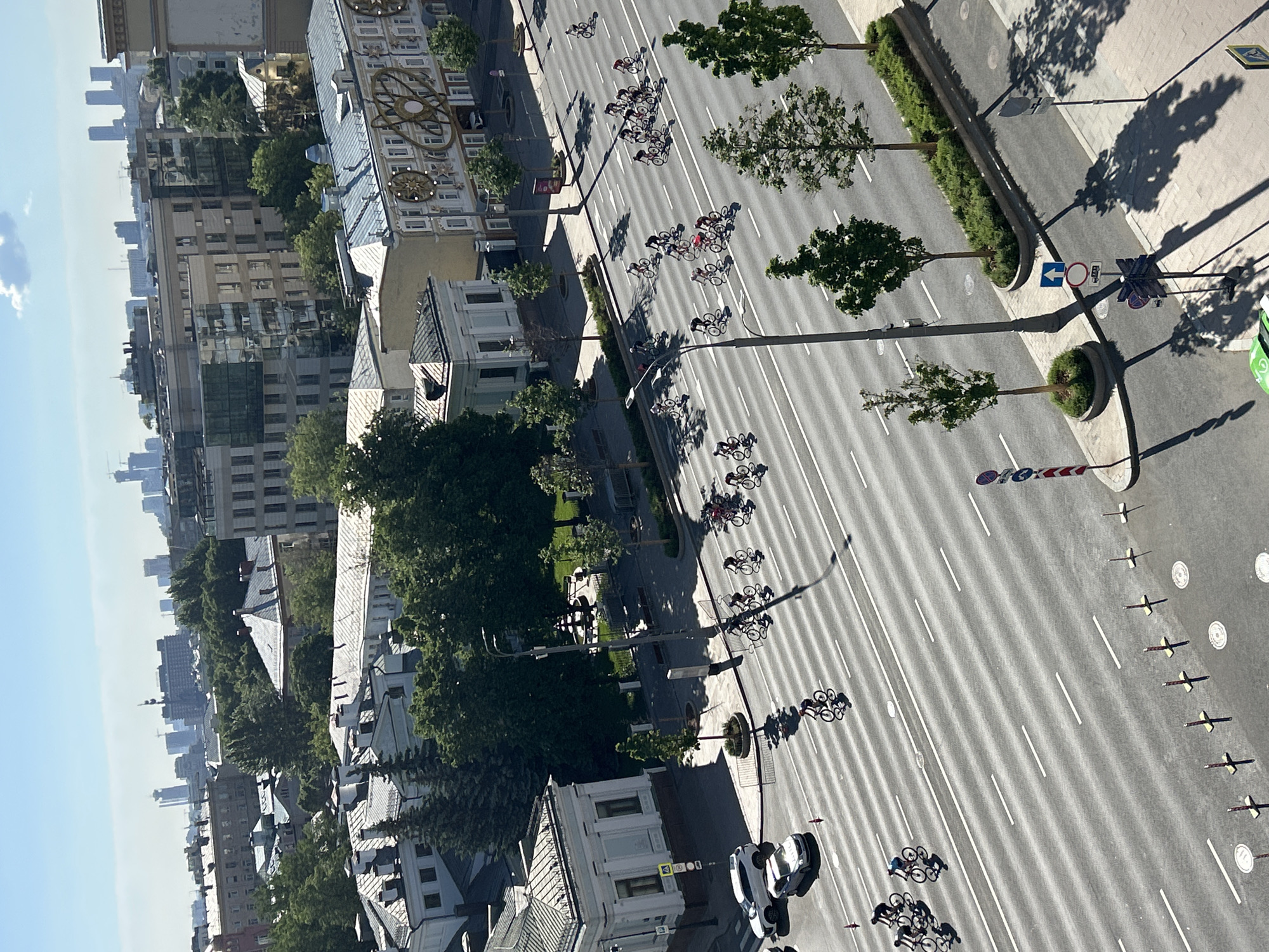 The image shows a street in a city, possibly Moscow, as seen from a high angle. A group of cyclists is moving along the road, which is lined with trees and buildings on both sides. The architecture of the buildings suggests a European style. There are several cars on the road as well, and street signs and markings are visible. In the distance, taller buildings can be seen, indicating a metropolitan area. The sky is clear, suggesting a sunny day.