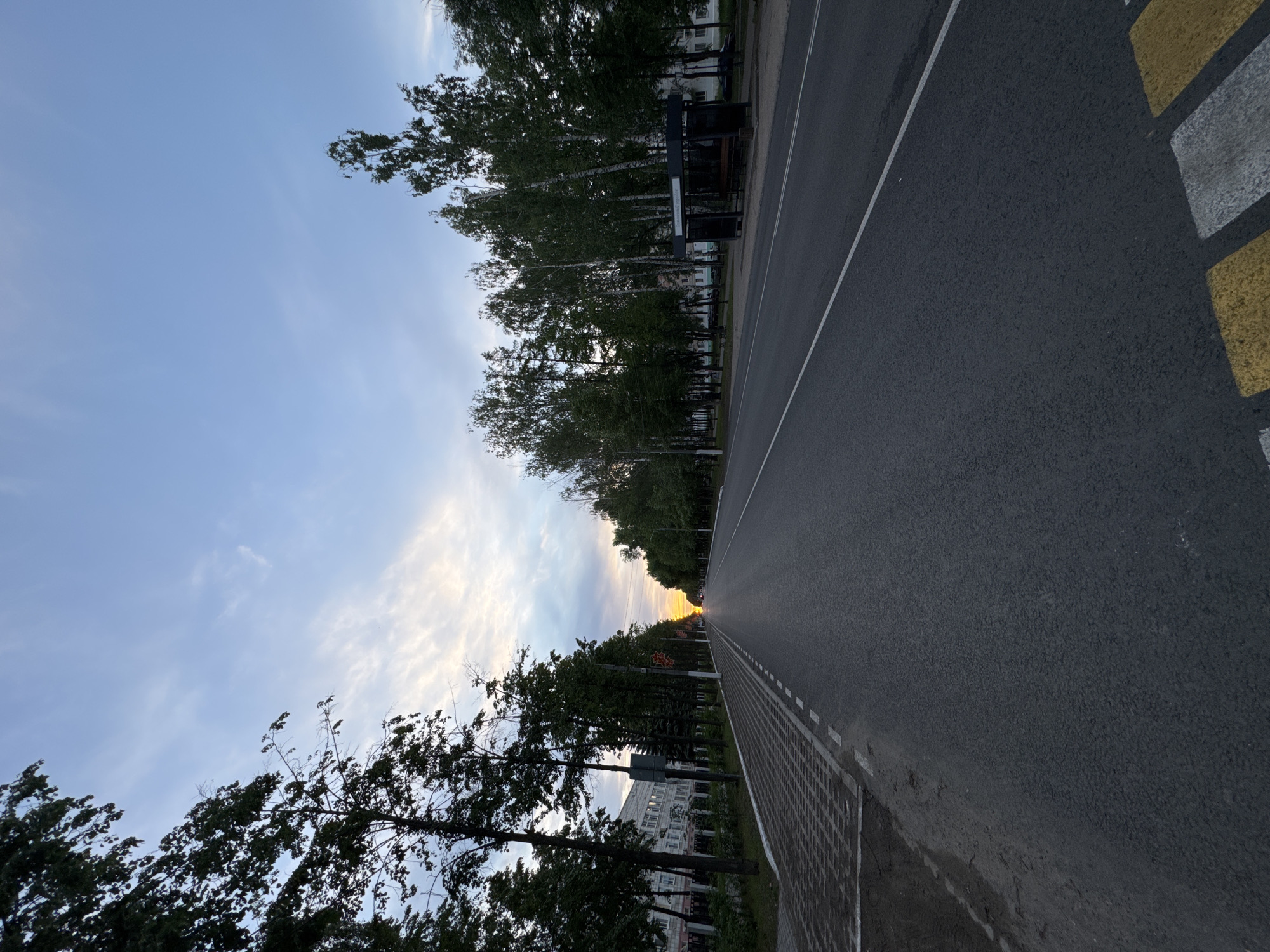 The image depicts a straight asphalt road in Chaikovsky, Russia, flanked by rows of trees. The road has a white line dividing the lanes and is marked with yellow and white zebra stripes on the far right. A bus stop is visible on the left side of the road, nestled among the trees. Buildings are partially visible beyond the trees on both sides of the road. The sky is a mix of blue and cloudy, with a hint of sunset glow in the distance at the end of the road. There are no people visible in the image.