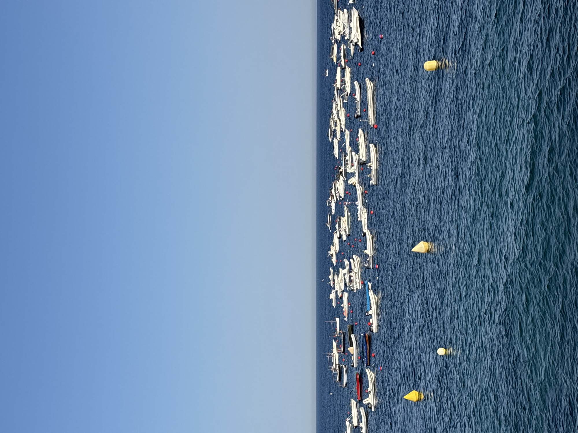 The image shows a wide view of the sea in Calella De Palafrugell, Spain, with a large number of small boats anchored in the water. The sky is clear and blue. The boats are mostly white, with some red and blue accents, and are arranged in multiple rows stretching across the frame. In the foreground, there are three buoys; two yellow and one white. The water is a deep blue with gentle ripples. The overall scene depicts a calm day at sea with numerous vessels moored offshore.