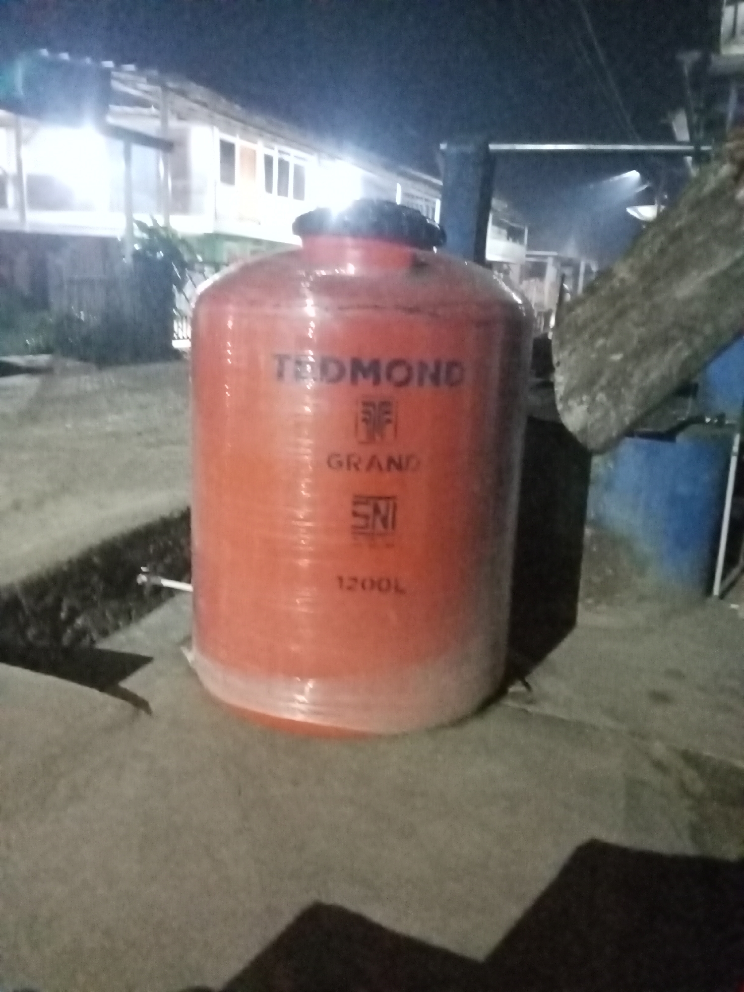 In Setia Jaya, Indonesia, at night, an orange water tank with a capacity of 1200 liters stands on a paved surface. The tank, branded "TEDMOND GRAND SNI," is partially wrapped in plastic. In the background, a building with multiple windows is illuminated, and a portion of a blue cylindrical object is visible to the right of the tank. The scene is dimly lit, suggesting it is outdoors.