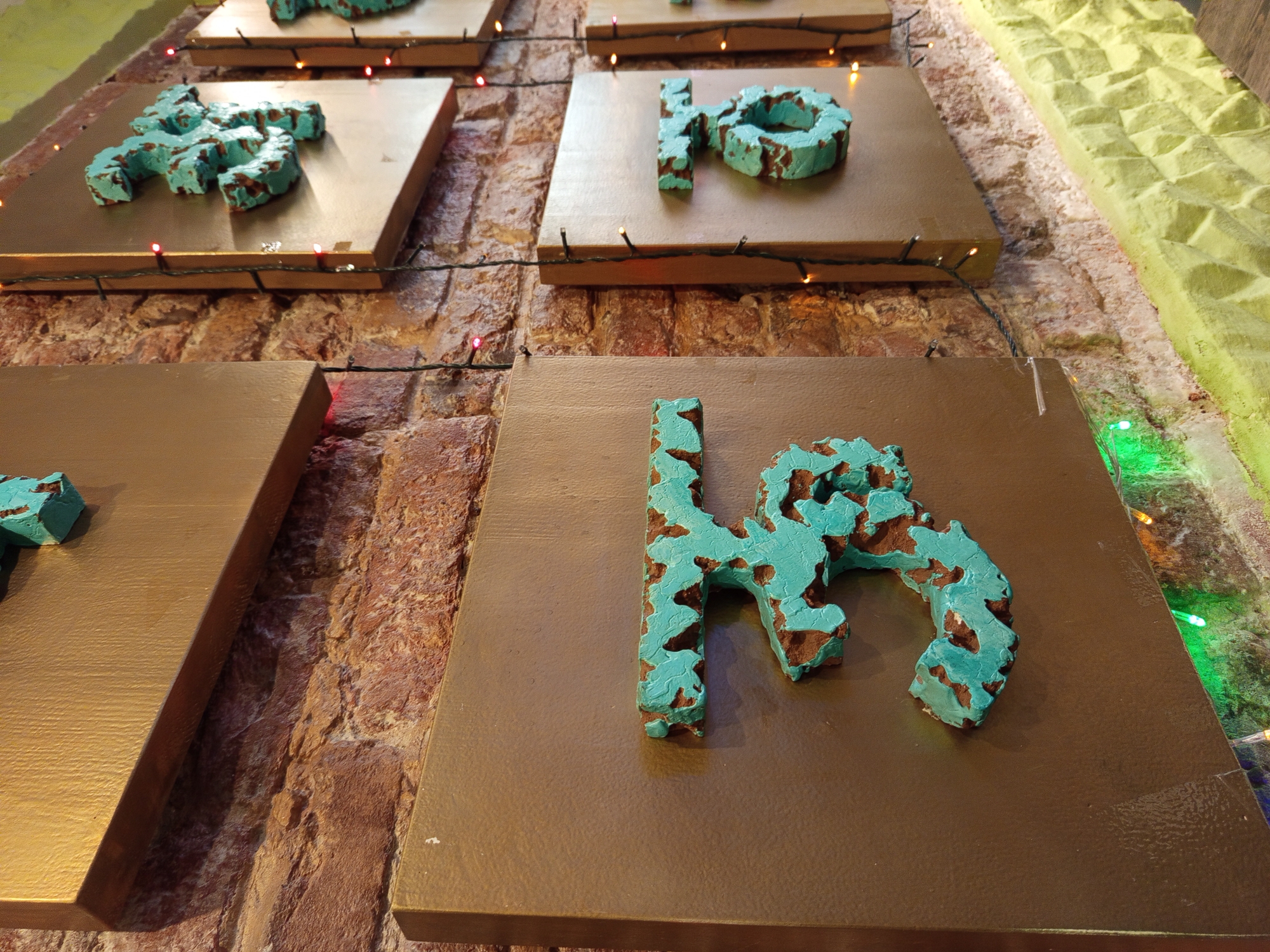 The image shows a close-up, eye-level view of decorative numbers mounted on a brick wall in a dimly lit establishment, possibly a bar or restaurant, in Saint Petersburg, Russia. The numbers, which appear to be "3" and "5", are crafted from a textured, turquoise-colored material with a distressed, aged look, mounted on square, brown backing panels. Red and green string lights are draped around the objects, with some lights casting a subtle glow on the wall and the lower part of the image. The brick wall has a rustic, reddish-brown hue, and the lower section of the wall is painted a vibrant yellow with a textured pattern. The perspective suggests the objects are part of a decorative display, potentially numbering tables or sections. No people are visible in the image. The overall ambiance is warm and inviting, with a hint of vintage charm.