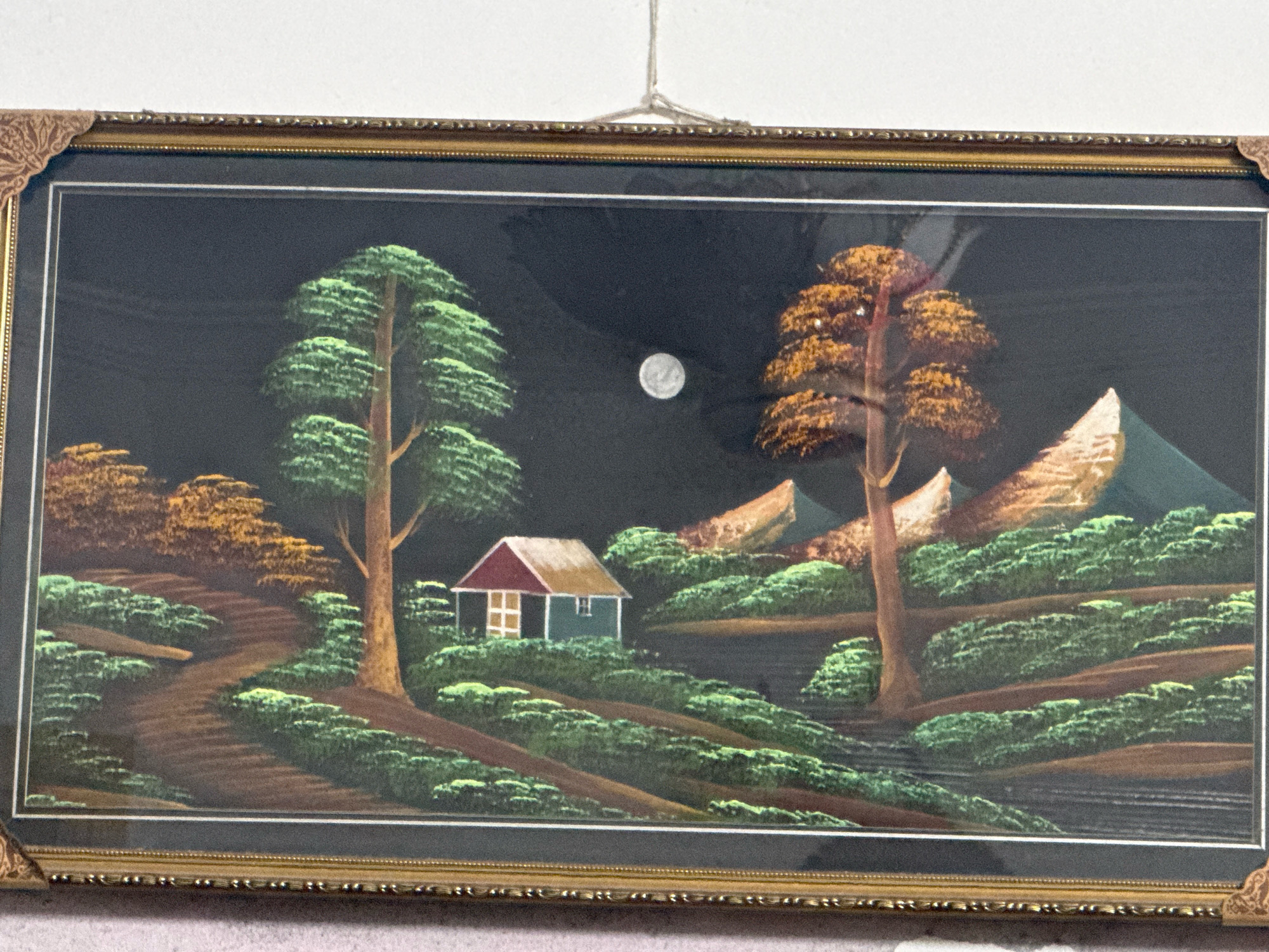 The image is a framed painting of a rural landscape at night. A full moon is visible in the dark sky. In the foreground, a winding path leads through rolling hills covered in green foliage. To the left, a tall evergreen tree stands beside the path. Further into the scene, a small house with a red roof and blue walls is nestled among the hills. To the right, a tree with orange foliage is prominent. In the background, mountains rise against the dark sky. The painting has a gold-colored frame with decorative corner accents. The painting's location in the city of Gujrat, Pakistan is not specified.