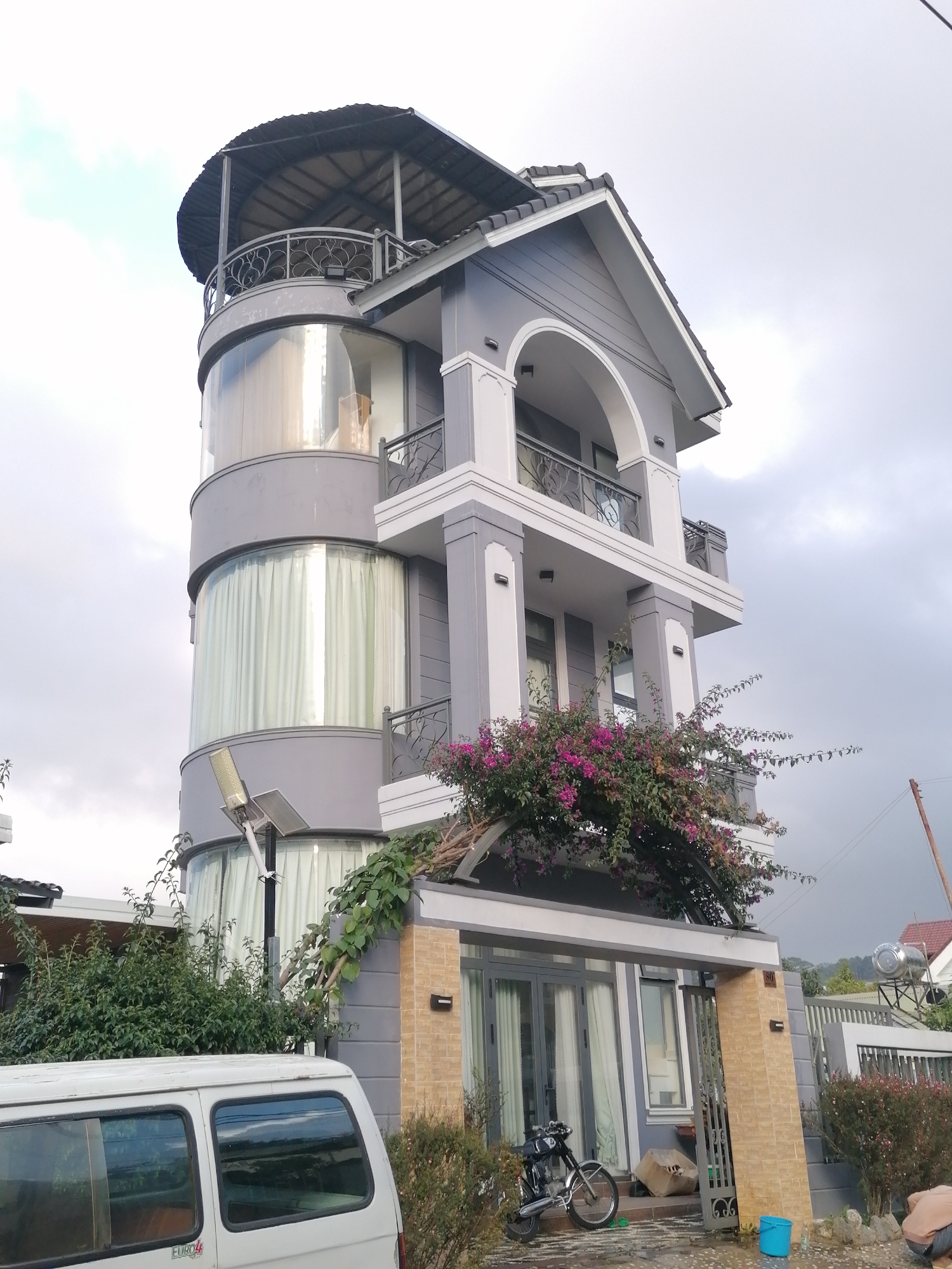 This is a full outdoor, eye-level shot of a tall, modern, multi-story building in the city of Xuân Trường - Đà Lạt, Vietnam. The building is painted in a cool gray color with white trim and features a rounded, glass-enclosed section on the left side, extending several stories high. The top of the building has a dark, curved roof supported by pillars, with a decorative metal railing on the balcony below.
On the right side of the building, there are balconies with similar railings and an arched architectural element over a covered entrance. Vibrant pink bougainvillea flowers spill over the railings and climb the side of the building, adding a pop of color to the otherwise muted palette.
In the foreground, a white van is parked on the left, its rear visible. To the right of the building's entrance, there's a black motorcycle parked on a stone pathway. A bright blue bucket sits near the entrance, and a few boxes are stacked on the ground. The ground appears wet, suggesting recent rain. Lush green bushes and trees surround the property. The sky is overcast with gray clouds. There are no people visible performing any actions. The overall impression is that of a residential or commercial property in a tropical or subtropical climate.
