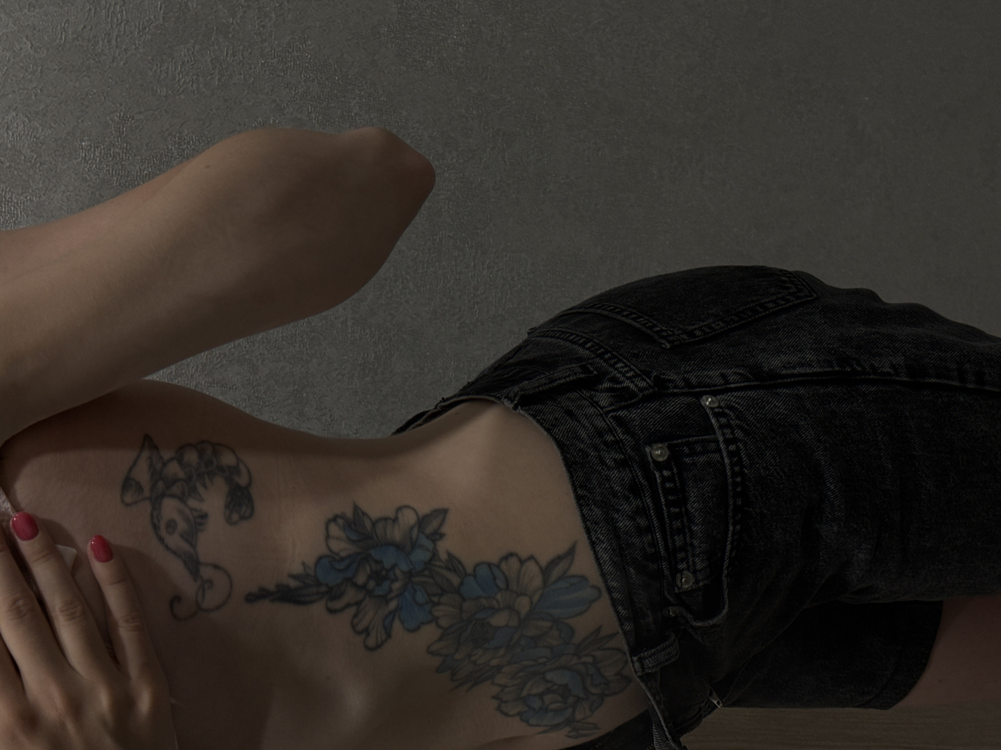 A side view shows the torso of a person from the waist up, against a grey textured wall. The person is wearing dark denim shorts. On the person's skin are two tattoos. The upper tattoo is a small, stylized depiction of a shrimp or crayfish on the ribcage. Below this, running down the side of the torso and wrapping around the hip, is a larger tattoo of blue and gray flowers with leaves. The person's hand, with red nail polish, is visible near the top of the frame, partially covering the upper tattoo. There is no identifiable location in Moscow, Russia.