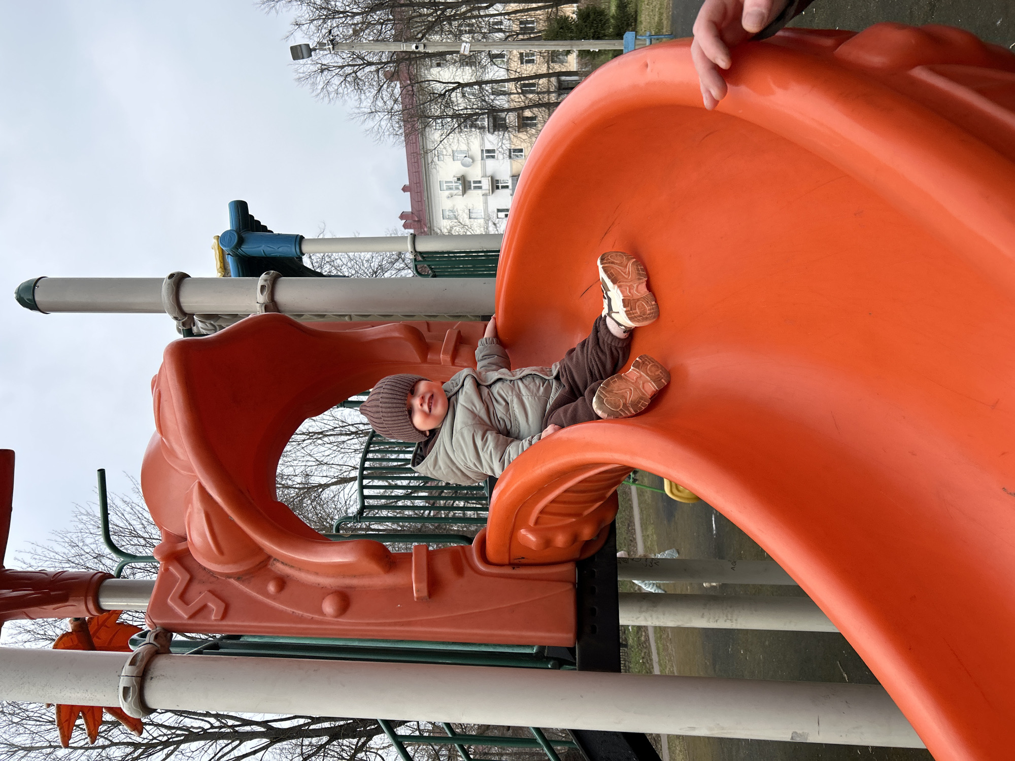A child is lying on a playground slide. The child is wearing a brown hat, a light grey jacket, and brown pants. The child is smiling. The slide is orange and part of a playground structure with white and green support bars. In the background, trees with bare branches are visible, along with a building with a red roof. This location appears to be in Klin, Russia. An adult's hand is partially visible at the top right of the image.