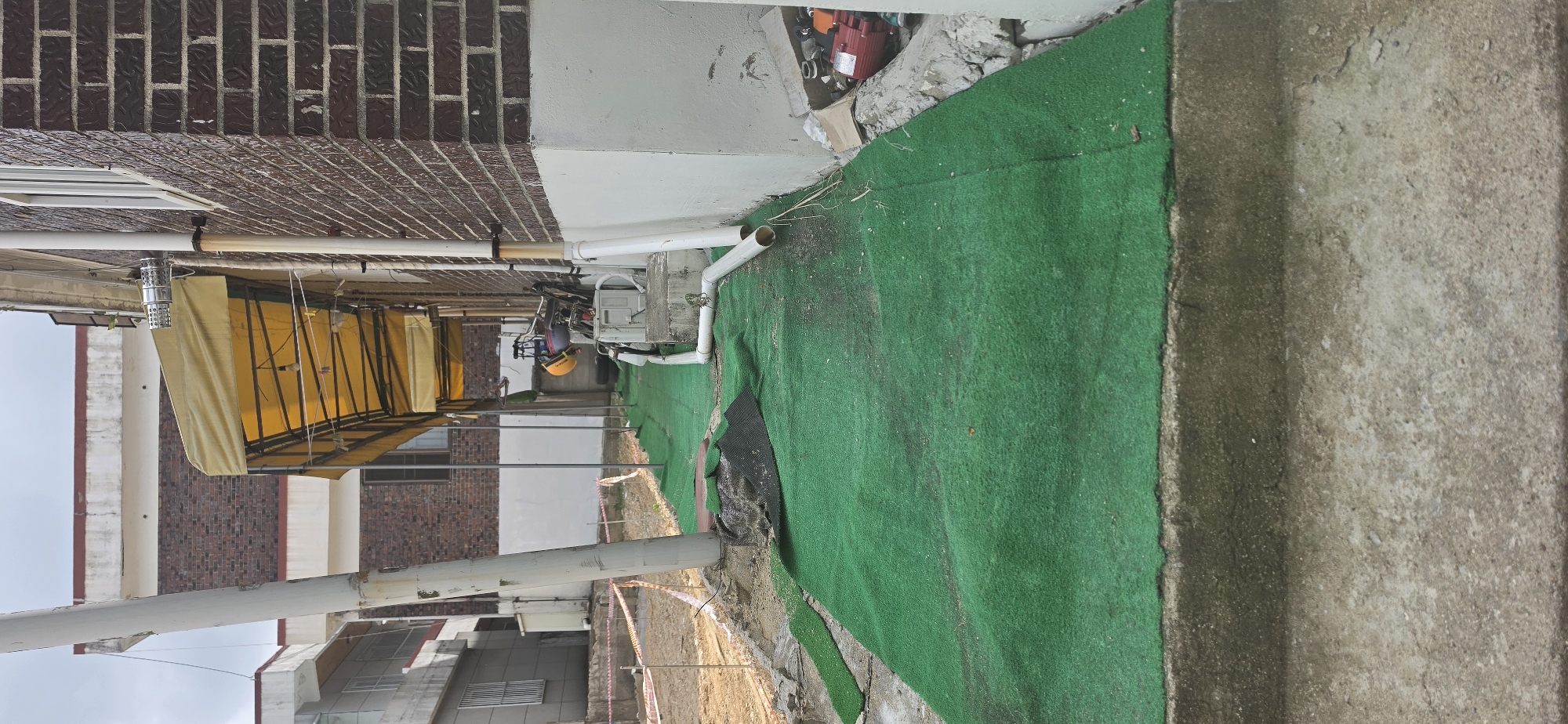 The image depicts a narrow, paved walkway covered with artificial green turf, running alongside a brick building. The walkway appears to be in a state of disrepair, with portions of the turf torn and dislodged. Construction fencing is visible in the background, suggesting ongoing work or maintenance in the area. Several PVC pipes are installed along the side of the building, some of which are exposed and disconnected. Various construction materials and equipment, including a red pump and a white unit, are scattered near the building's foundation. The walkway leads towards a building with a yellow awning, further down the path. The overall scene suggests an outdoor area in Cheongju-si, South Korea, possibly a commercial or residential complex undergoing renovation or a neglected outdoor space.