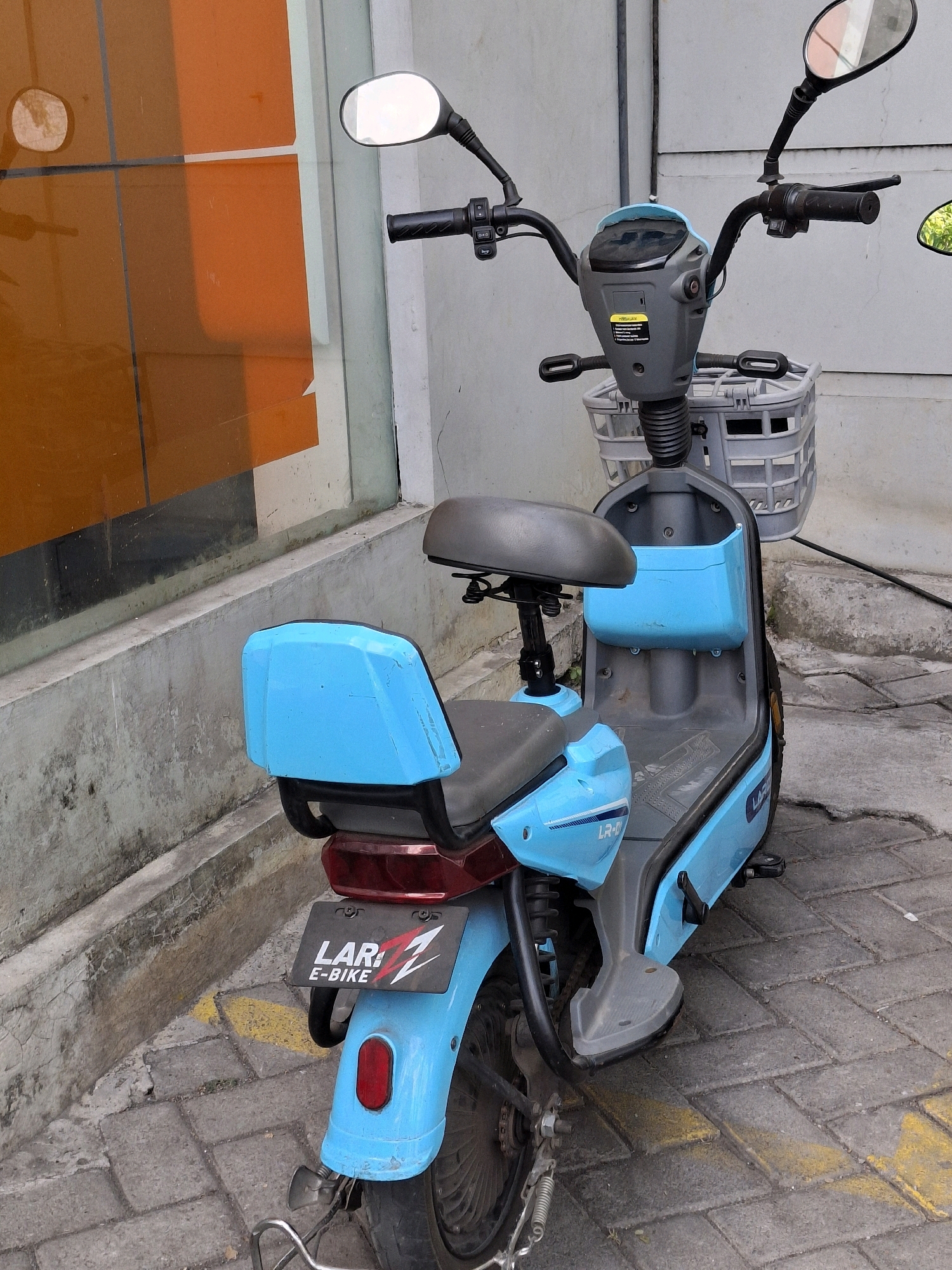 An outdoor medium shot captures a blue electric bicycle parked on a paved surface near an orange-paneled wall. The bicycle, oriented slightly towards the viewer's right, is the primary subject. It features a grey handlebar with mirrors and controls, a grey central body, a grey seat, and a blue rear seat. A grey basket is attached to the front of the bicycle. A license plate with "LAR 1 E-BIKE" is affixed to the rear fender. The ground is composed of grey interlocking bricks with yellow painted markings. The background consists of a textured concrete wall and an orange paneled building. The scene is devoid of any people. The lighting suggests it is daytime.