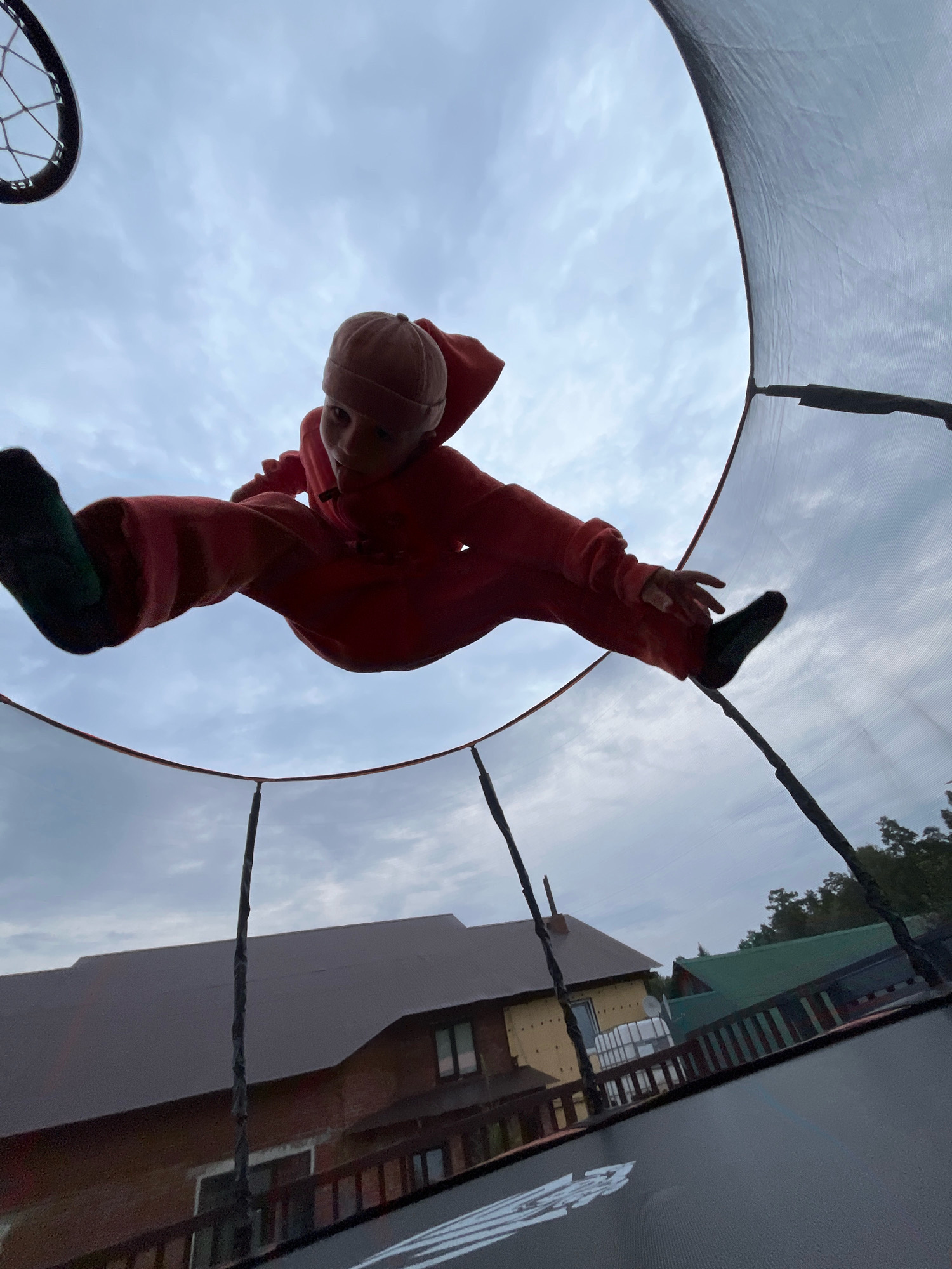 An upward-angle, low-light shot captures a child in mid-air on a trampoline. The child, wearing a red jumpsuit and a light-colored beanie, is performing a straddle jump, with legs extended to the sides. The background is a cloudy sky. Below the trampoline, houses with dark brown and green roofs, and trees are visible. The scene appears to be in a suburban or rural area.
