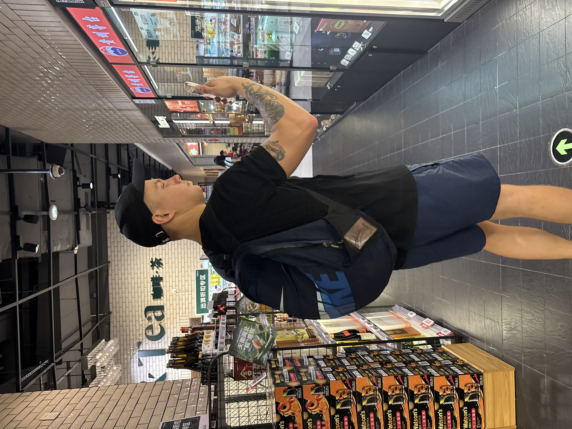 A young Caucasian male wearing a black baseball cap, black t-shirt, and navy blue shorts stands in a grocery store aisle, facing away from the camera. He is wearing a navy blue backpack with a large blue Nike logo on it. He is holding a clear smartphone in his right hand, appearing to take a picture or record video. The aisle is lined with shelves displaying various products. To the left, there are stacks of orange boxes labeled "Watson's". To the right, there are glass display cases with illuminated shelves showcasing bottled beverages and other items. A sign above the display cases reads "贵州茅台" (Guizhou Maotai). The store has a tiled floor and ceiling lights. The environment suggests a retail setting, likely a supermarket or convenience store in Haizhu District, China, as indicated by the signage.