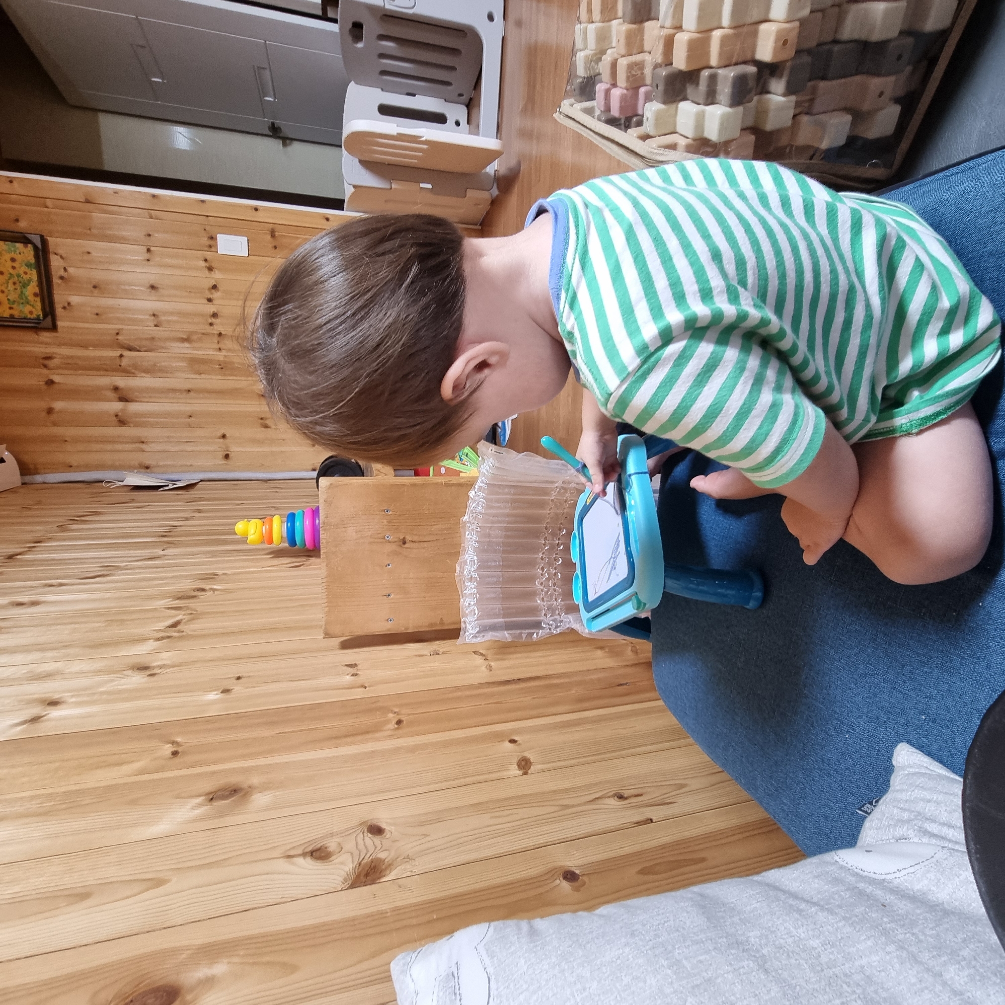 In a room with wood-paneled walls, a toddler sits cross-legged on a dark blue cushioned surface, facing a small aqua-colored drawing board with a stylus. The toddler, wearing a green and white striped t-shirt, is actively drawing on the board, with their right hand holding the stylus. Behind the drawing board, a plastic bubble wrap barrier is visible. To the left of the toddler, a portion of a light-colored cushion is visible. In the background, a stacking toy with colored rings and a yellow duck figure rests on a wooden shelf unit. Further in the background, a white baby gate is partially visible, and a wooden cabinet with visible screws is present. The flooring is a light brown wood. The scene appears to be indoors, possibly a home, in Dunnae-myeon, South Korea, given the context.