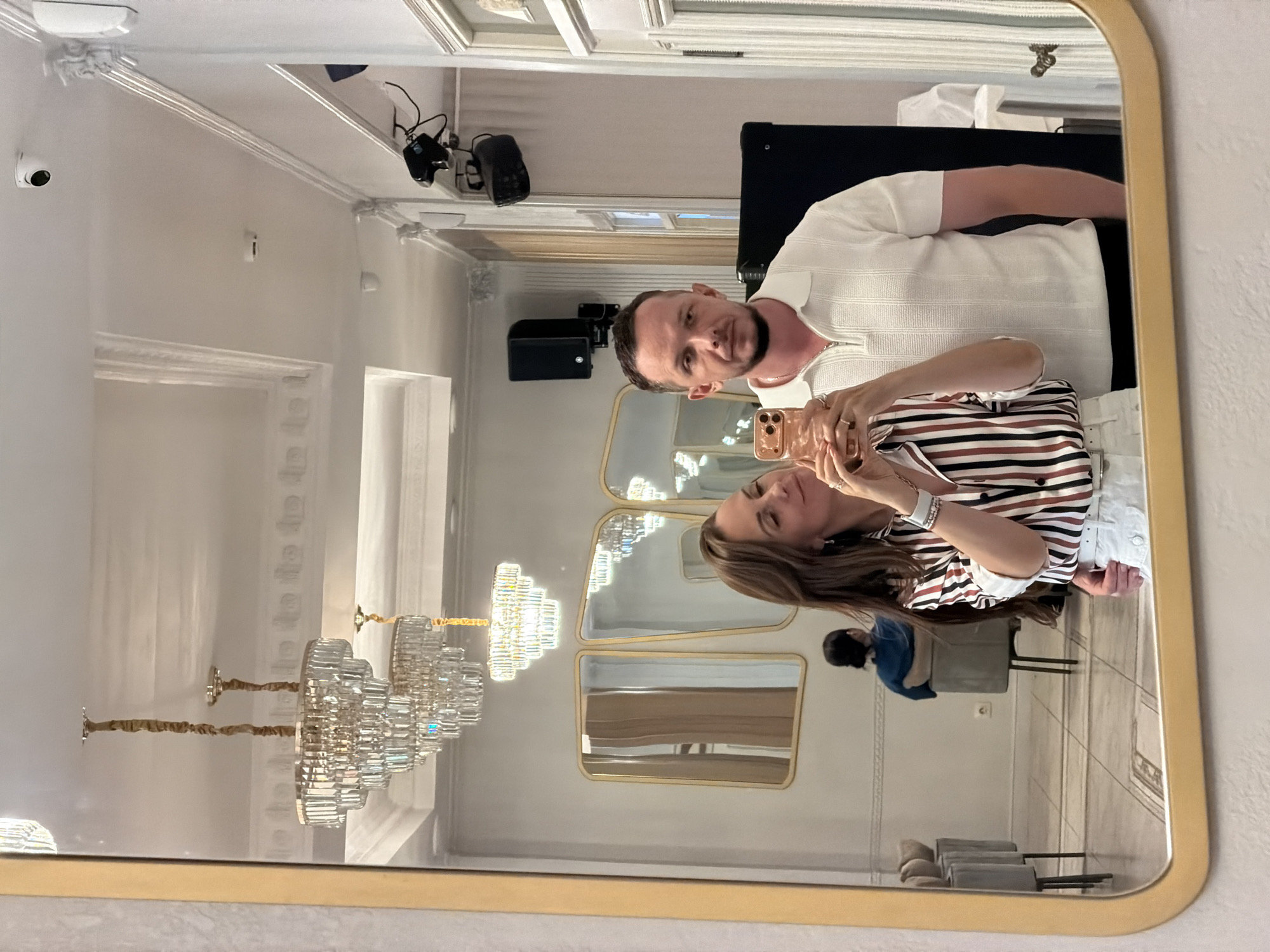 A couple is taking a selfie in a mirror in a well-lit, upscale interior that appears to be a restaurant or lounge. The woman, on the left, is wearing a striped shirt and white shorts, holding a rose-gold smartphone to take the photo. The man, on the right, is wearing a white polo shirt and black pants, looking directly at the camera. In the background, reflected in the mirror, are ornate chandeliers, decorative molding on the ceiling, framed mirrors on the wall, and seating areas with chairs and tables. A person is visible sitting in a chair in the middle ground, with their back to the camera. The scene is clean and modern with a touch of classic elegance. The location in Birobidzhan, Russia cannot be determined from the visual information provided in the image.