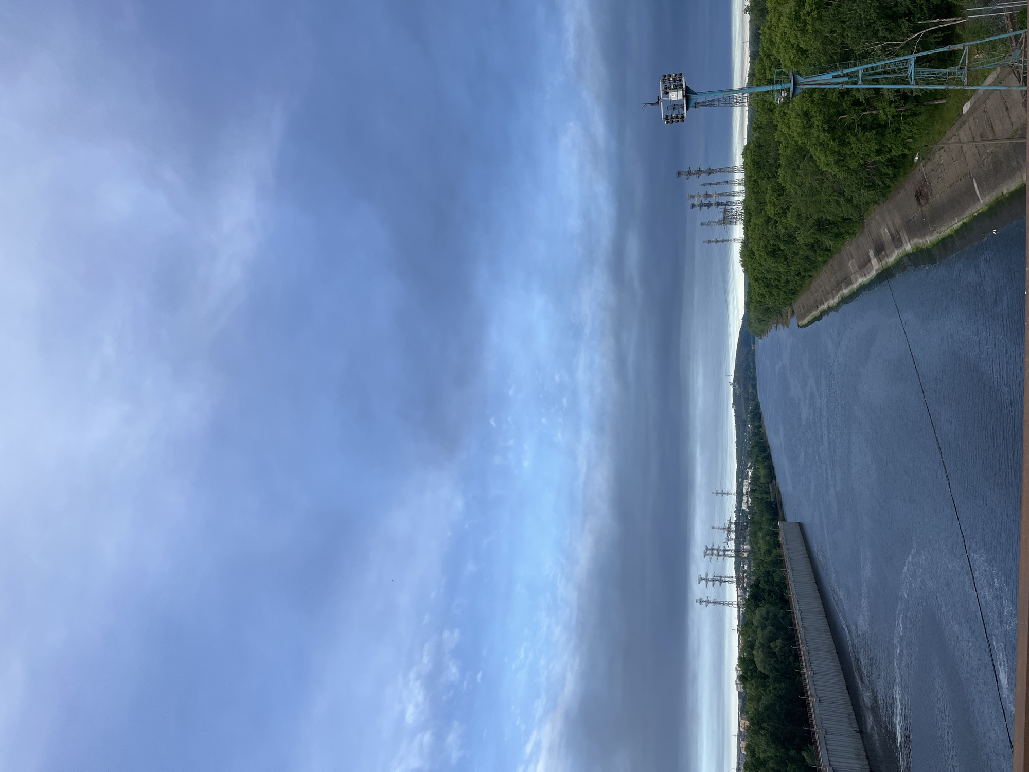 The image depicts a water body, likely a river or reservoir, adjacent to a forested bank. A concrete structure, possibly a dam or retaining wall, runs along the edge of the water. In the distance, power lines are visible near the forested area and a tower. The sky is predominantly blue with scattered clouds. Based on the location mentioned, the image likely shows the Votkinsk Reservoir in Chaikovsky, Russia.