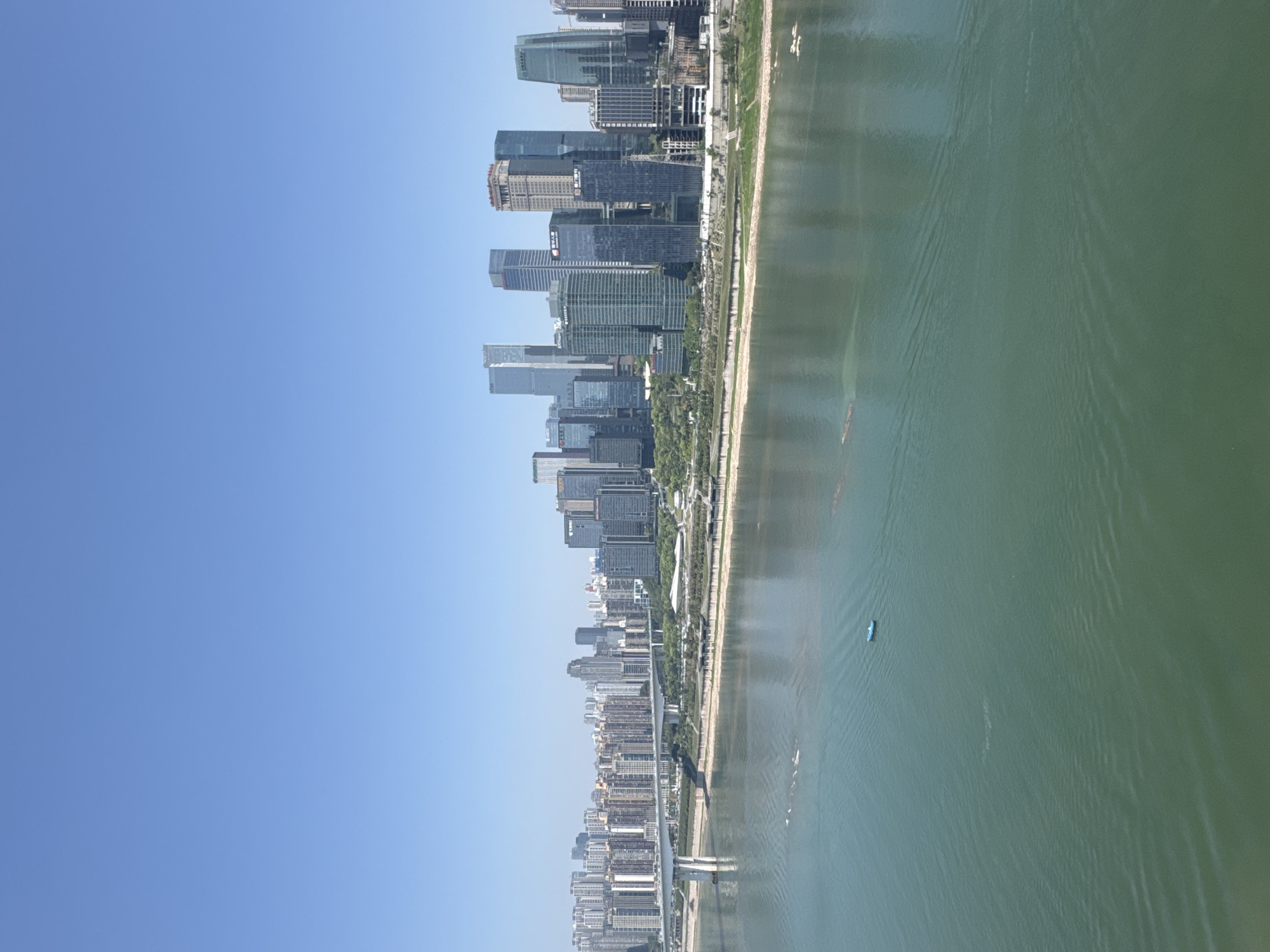 The image shows a cityscape in Yuzhong District, Chongqing, China, taken from an elevated perspective. On the left is a clear, blue sky. The right side of the image features a body of water with ripples, likely the Yangtze River. Along the shoreline, a line of trees and green space is visible, adjacent to which stands a dense collection of modern skyscrapers and buildings. A small blue boat is visible on the water.
